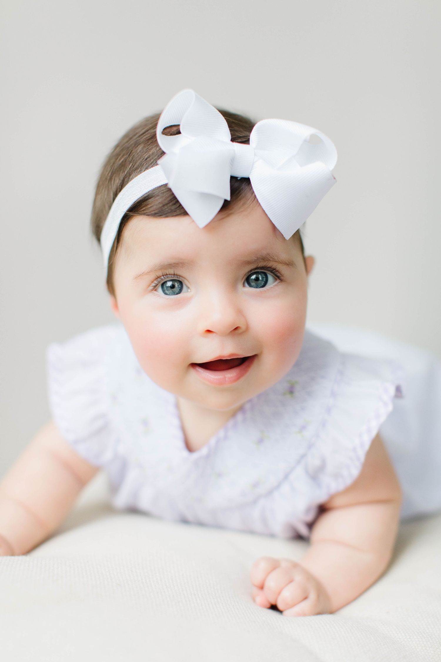 A baby wearing a white bow and dress smiles playfully while lying on their stomach across multiple photos.