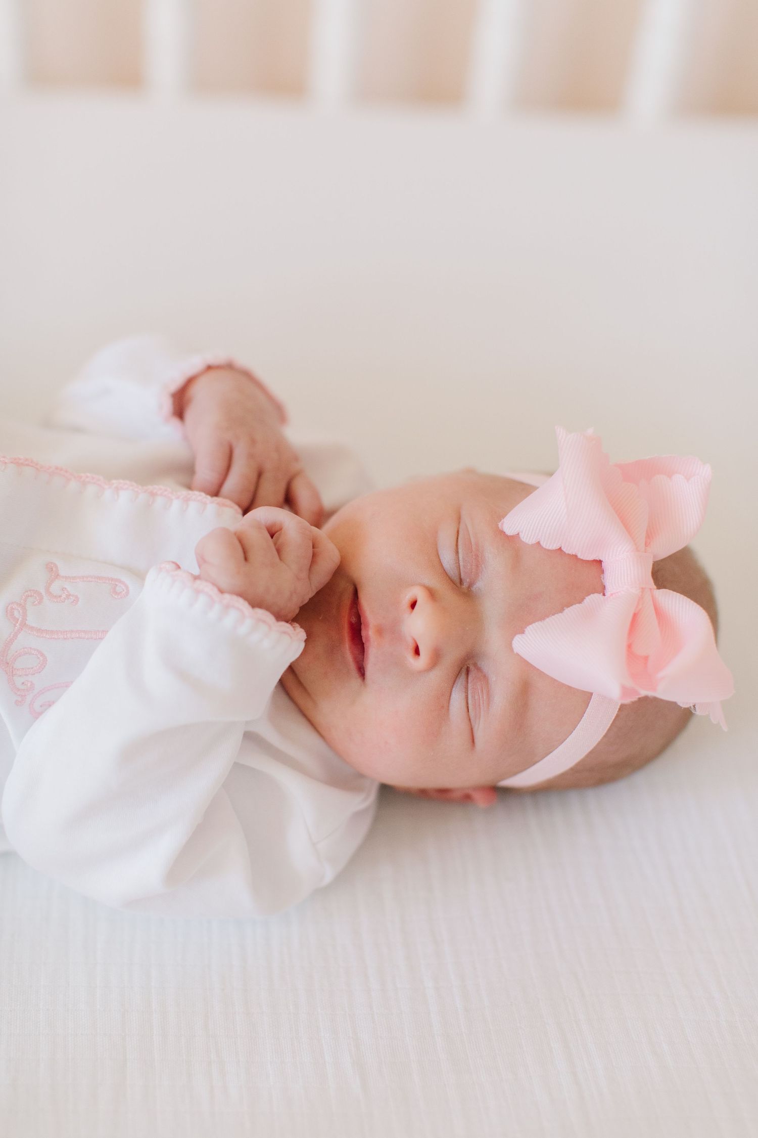 Sleeping newborn wearing white outfit and pink bow headband on white bedding.