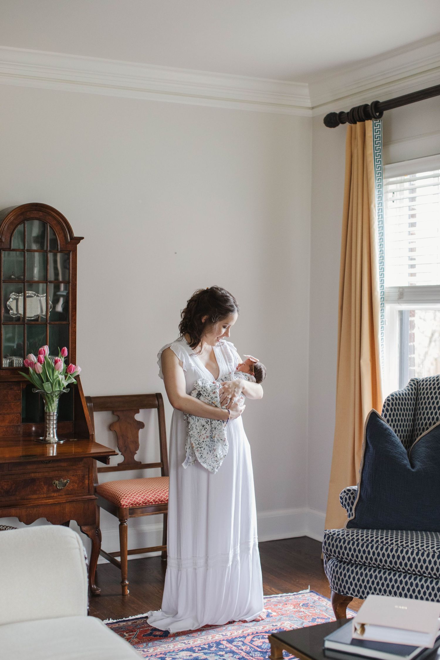 Parent in white dress holding baby in elegant living room with classic furniture.