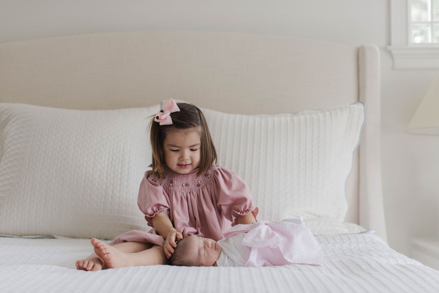 Little girl in pink dress sits on white bed with matching hair bow.