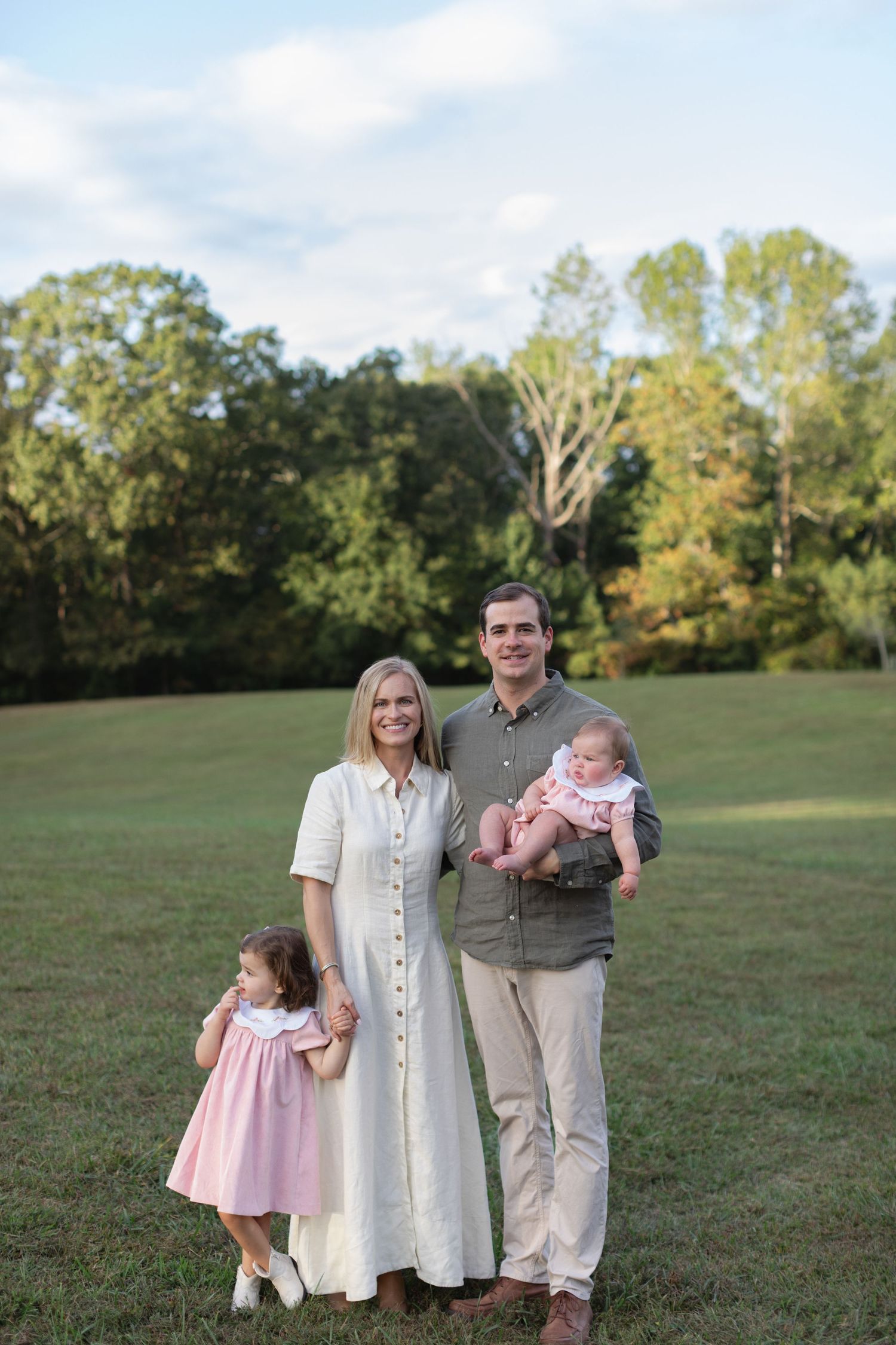 Family in coordinating neutral outfits poses together on grassy field during golden hour outdoor photoshoot.