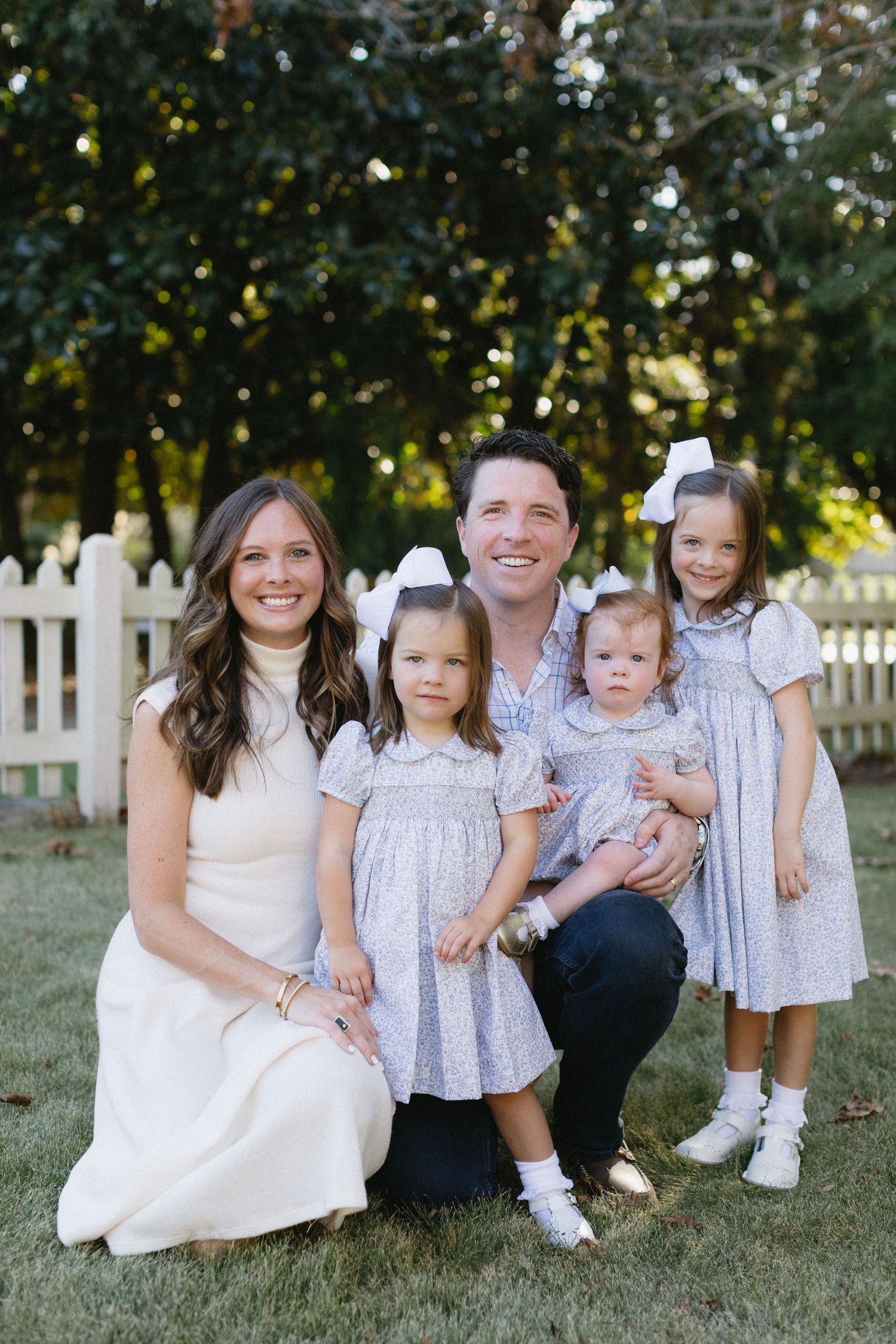 Family wearing coordinated white and blue outfits sits together on grass for outdoor portrait session.
