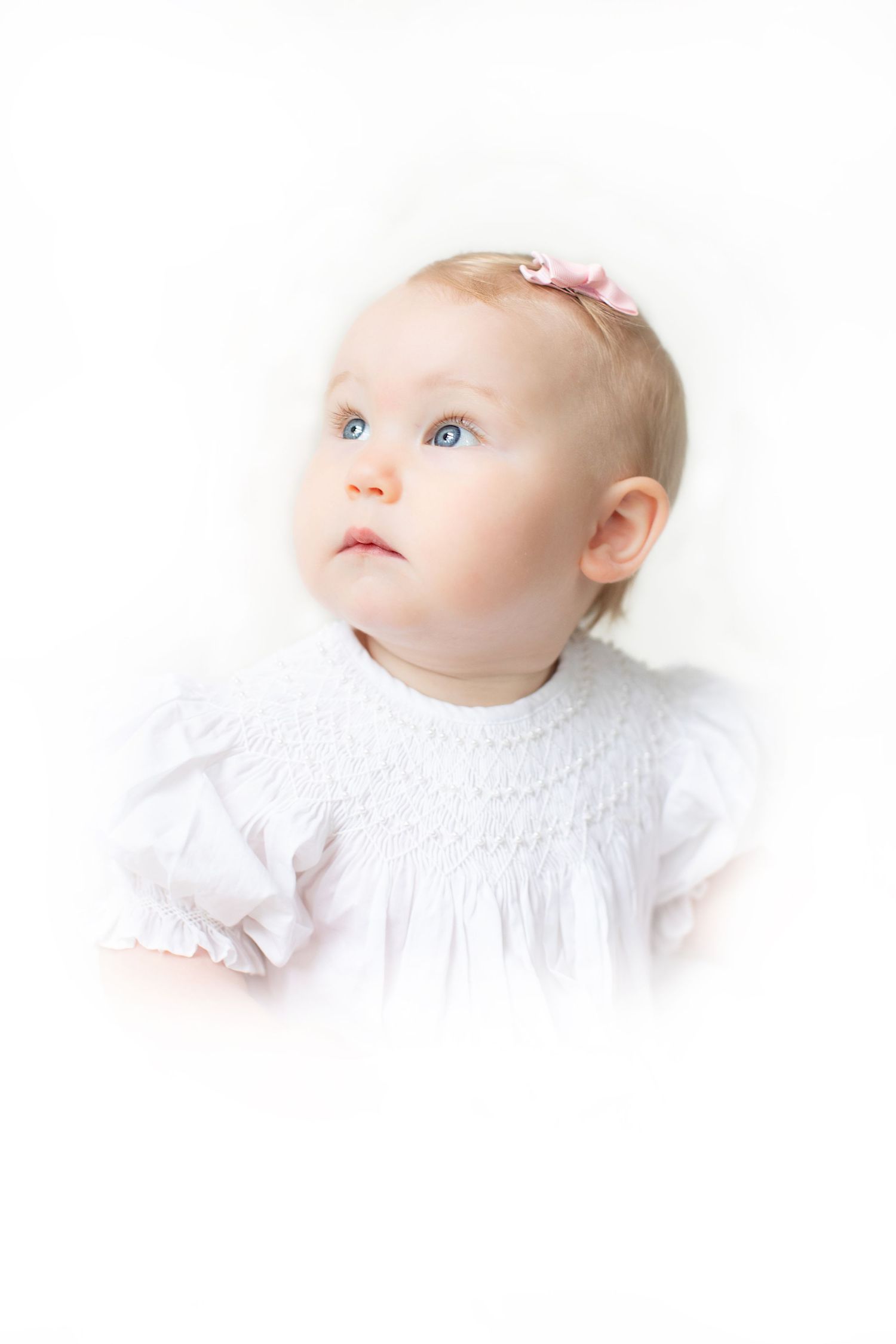 A blue-eyed baby girl in a white smocked dress with a pink bow headband looks upward against a white background.