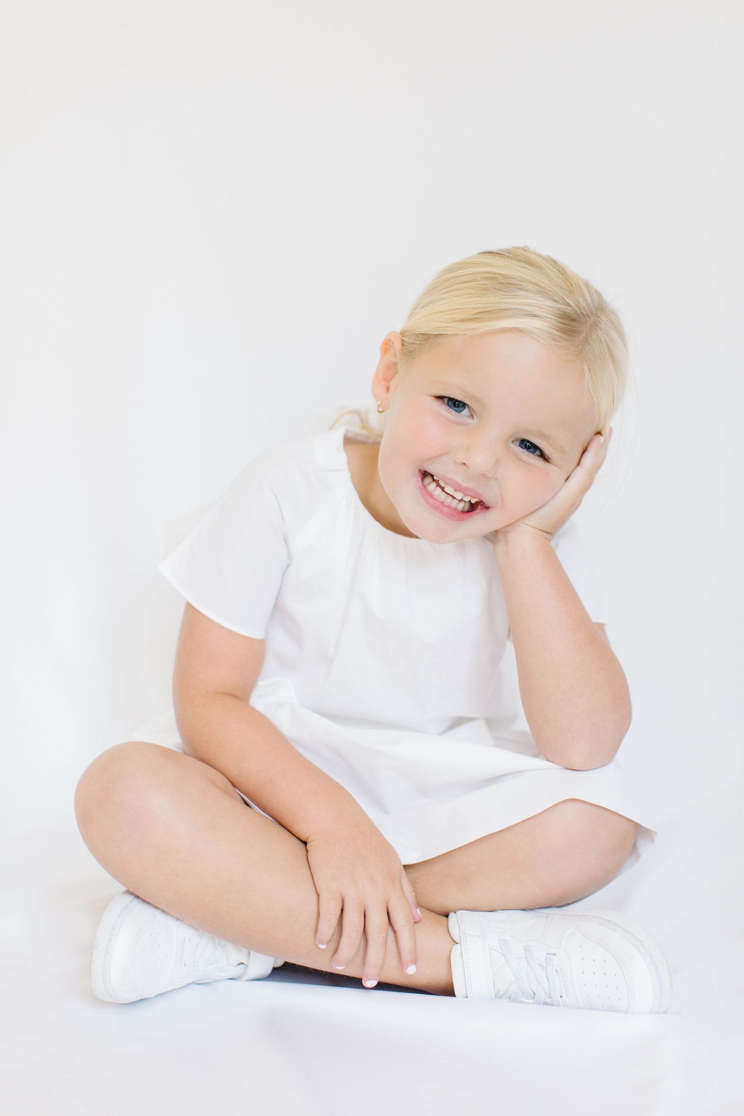 Child wearing white sitting cross-legged on white background displaying joyful expression and resting head on hand.