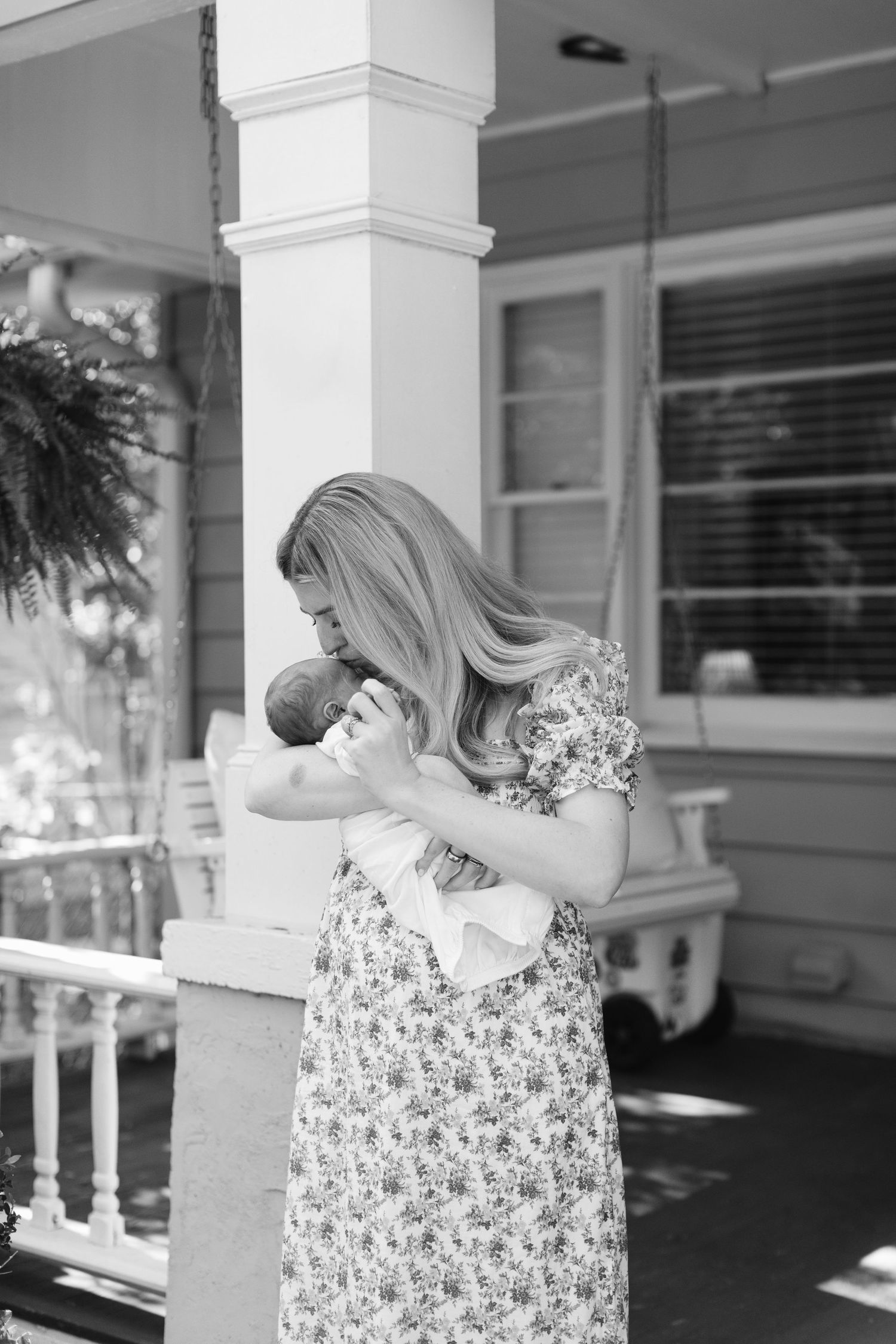 Black and white photo of a mother in a floral dress cradling her newborn baby on a classic Southern porch.