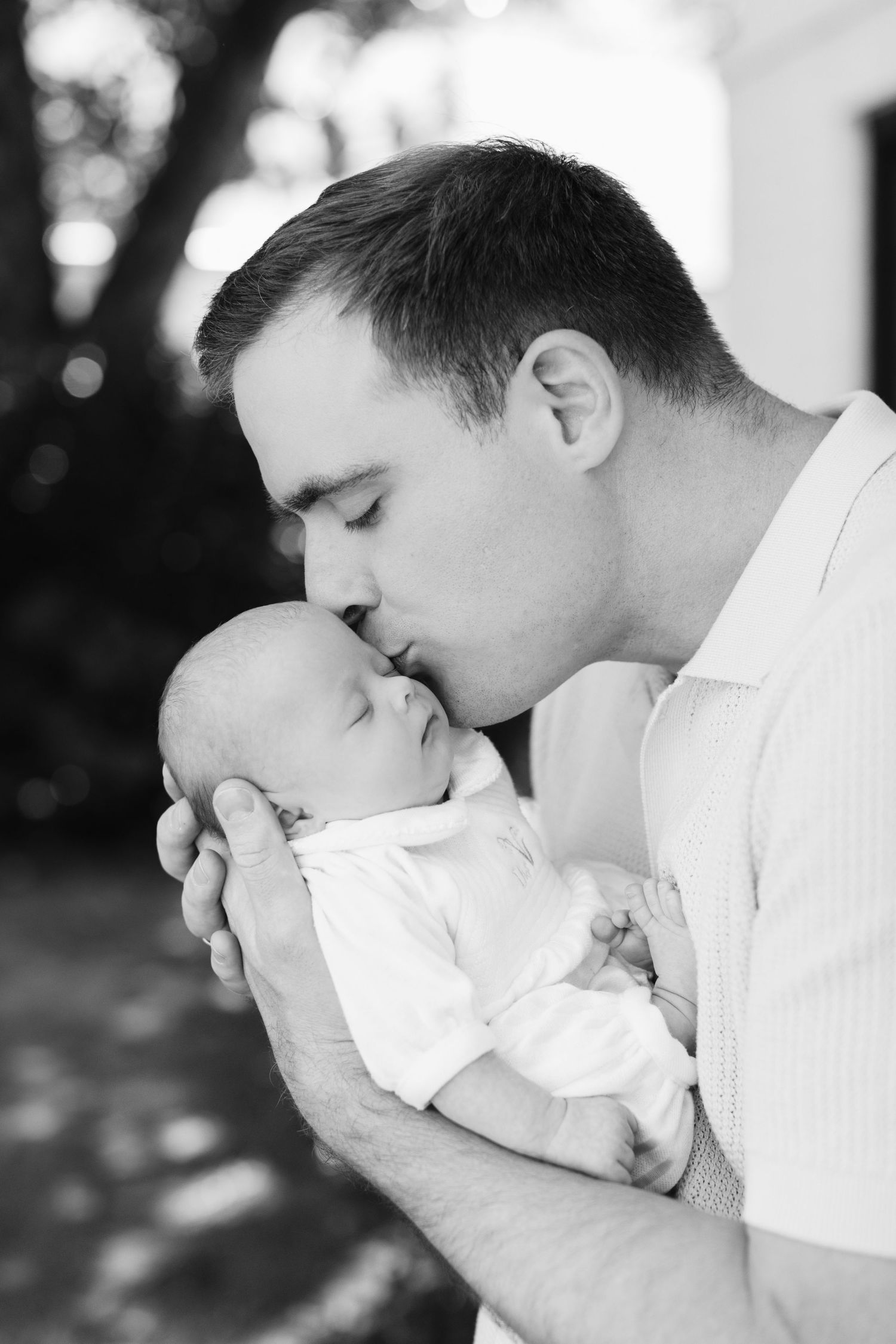 A father tenderly kisses his sleeping newborn baby in a black and white portrait photo.