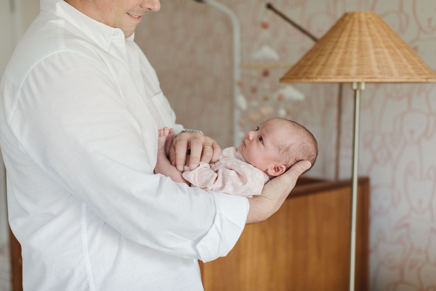 Parent cradling newborn baby in soft pink clothing near vintage table lamp.
