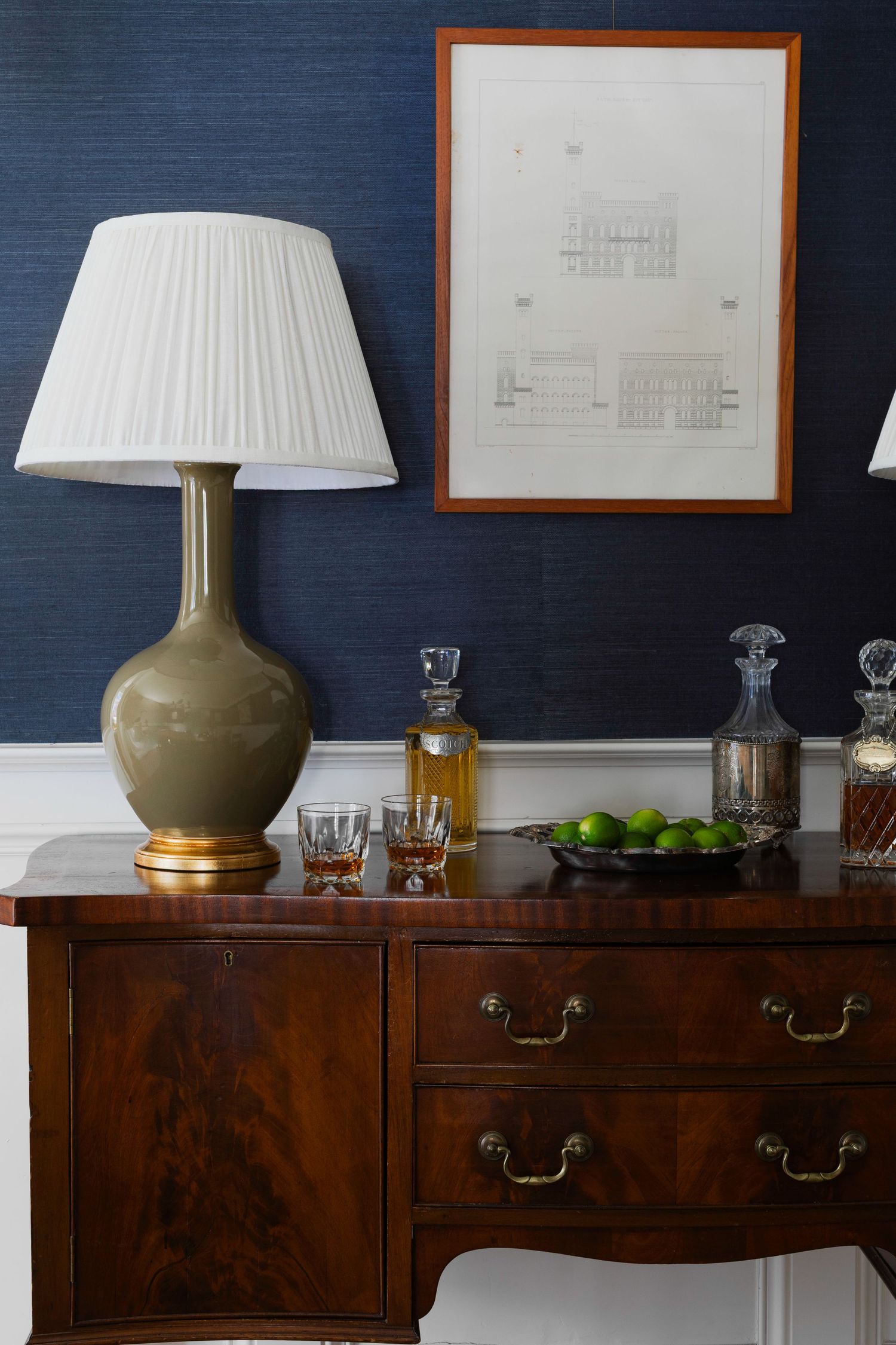 Olive green ceramic table lamp with white pleated shade sits atop dark wood sideboard against navy blue wall.