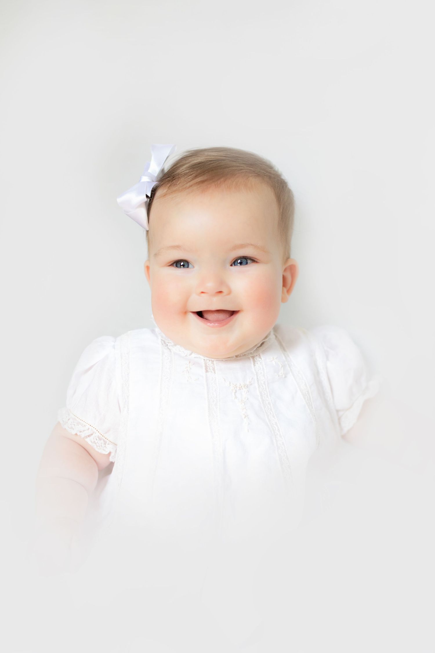 Smiling baby girl in white dress with bow hair clip lying on white background.
