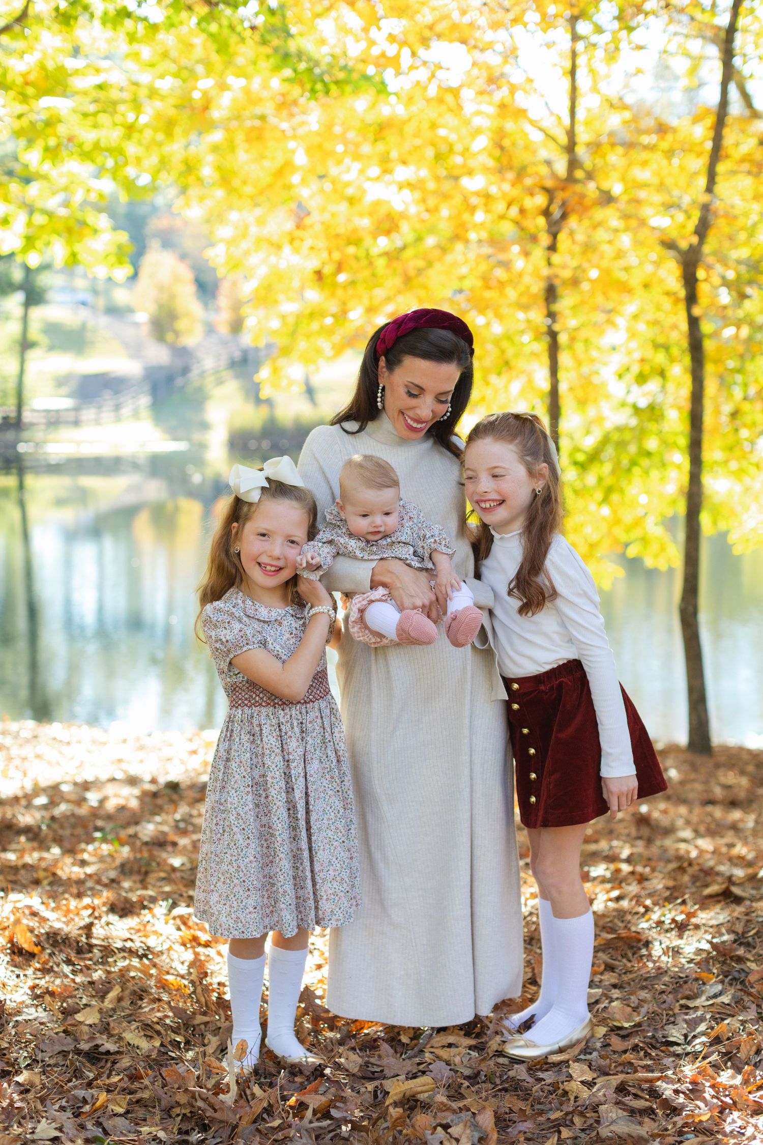 Three children in vintage-style dresses play together in autumn leaves with golden trees in the background.