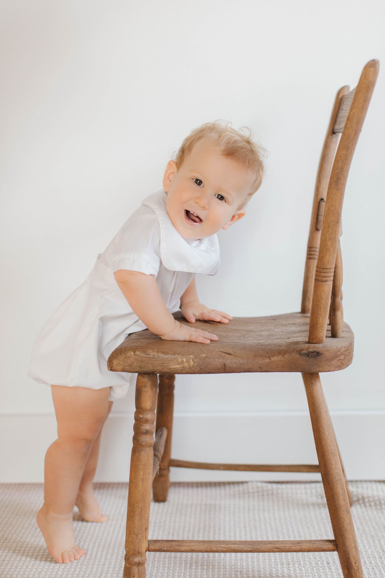 A smiling toddler in a white outfit stands holding onto a wooden chair on a light carpet.