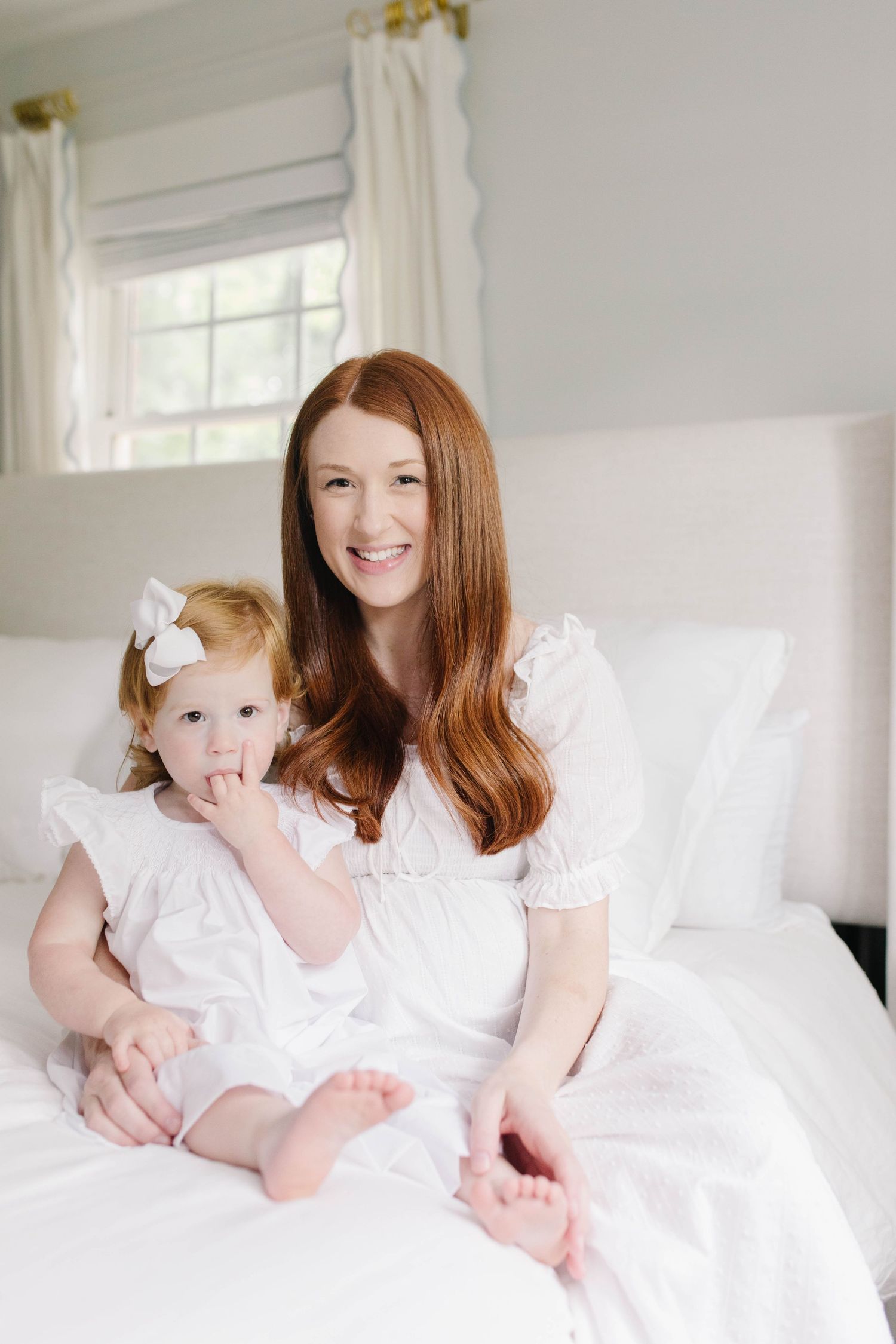 Mother and child in matching white outfits sit together on a bright bed near a window.