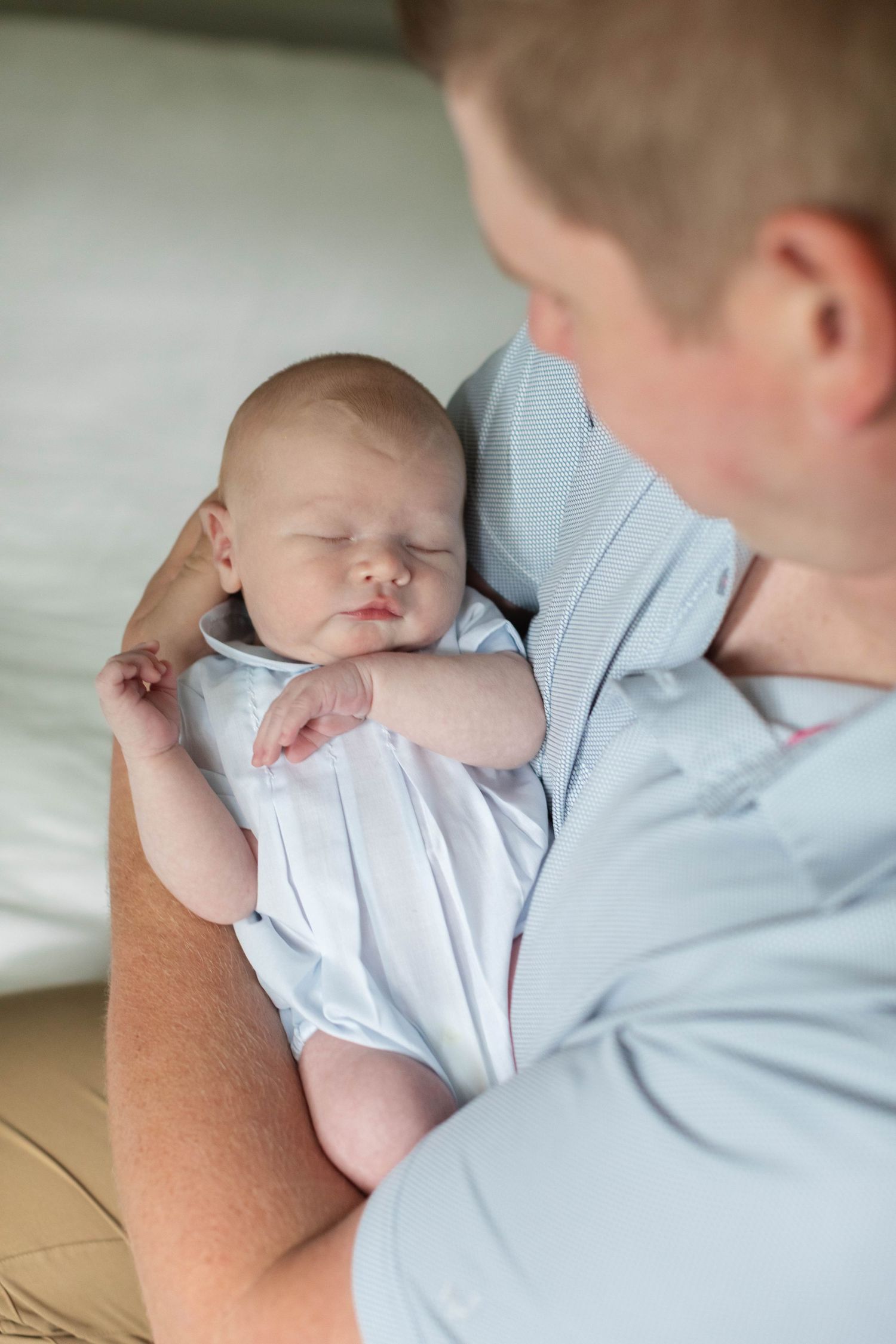 A sleeping newborn baby in white clothing rests peacefully against someone wearing a light blue shirt.