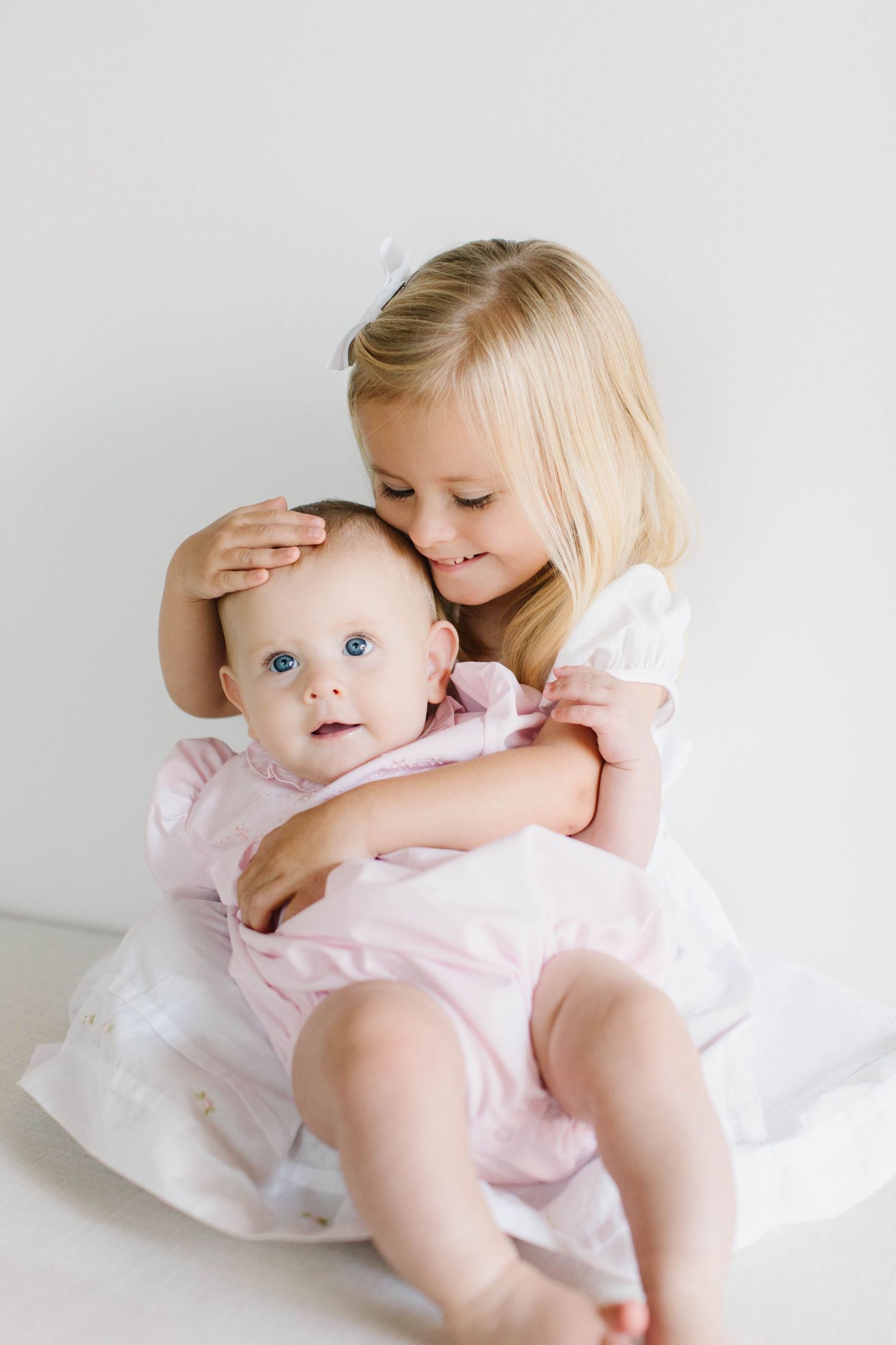 Two children in white and pink outfits share a tender moment against a light background.