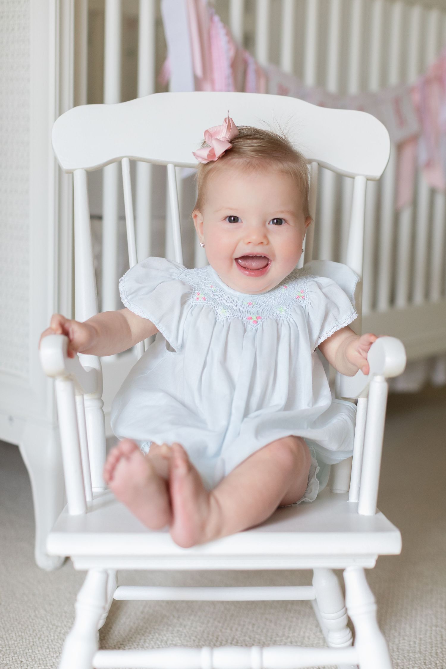 Happy baby girl in white smocked dress sitting in white rocking chair, smiling in bright nursery room.