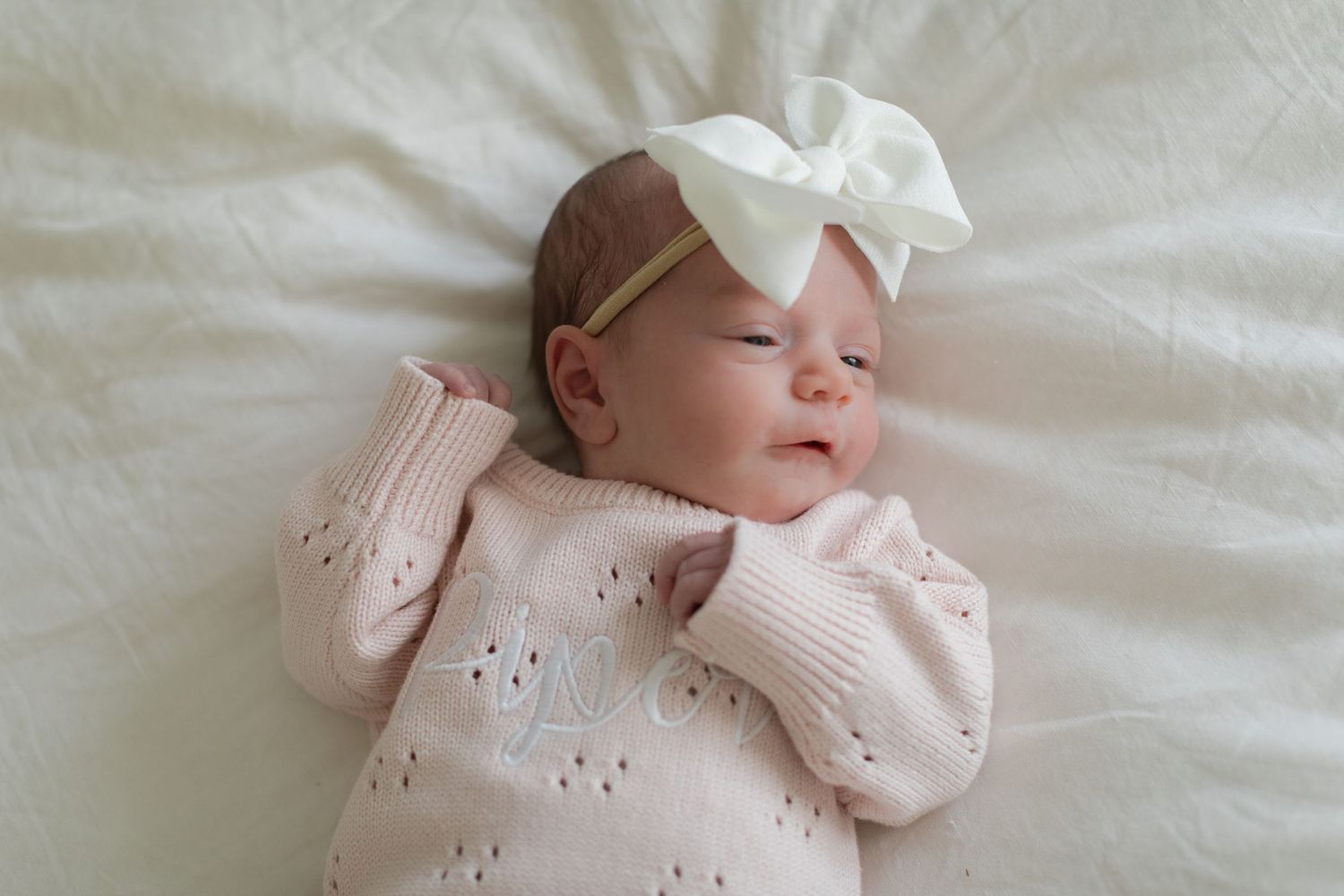 Newborn baby wearing a pink bow headband sleeps peacefully on white bedding.