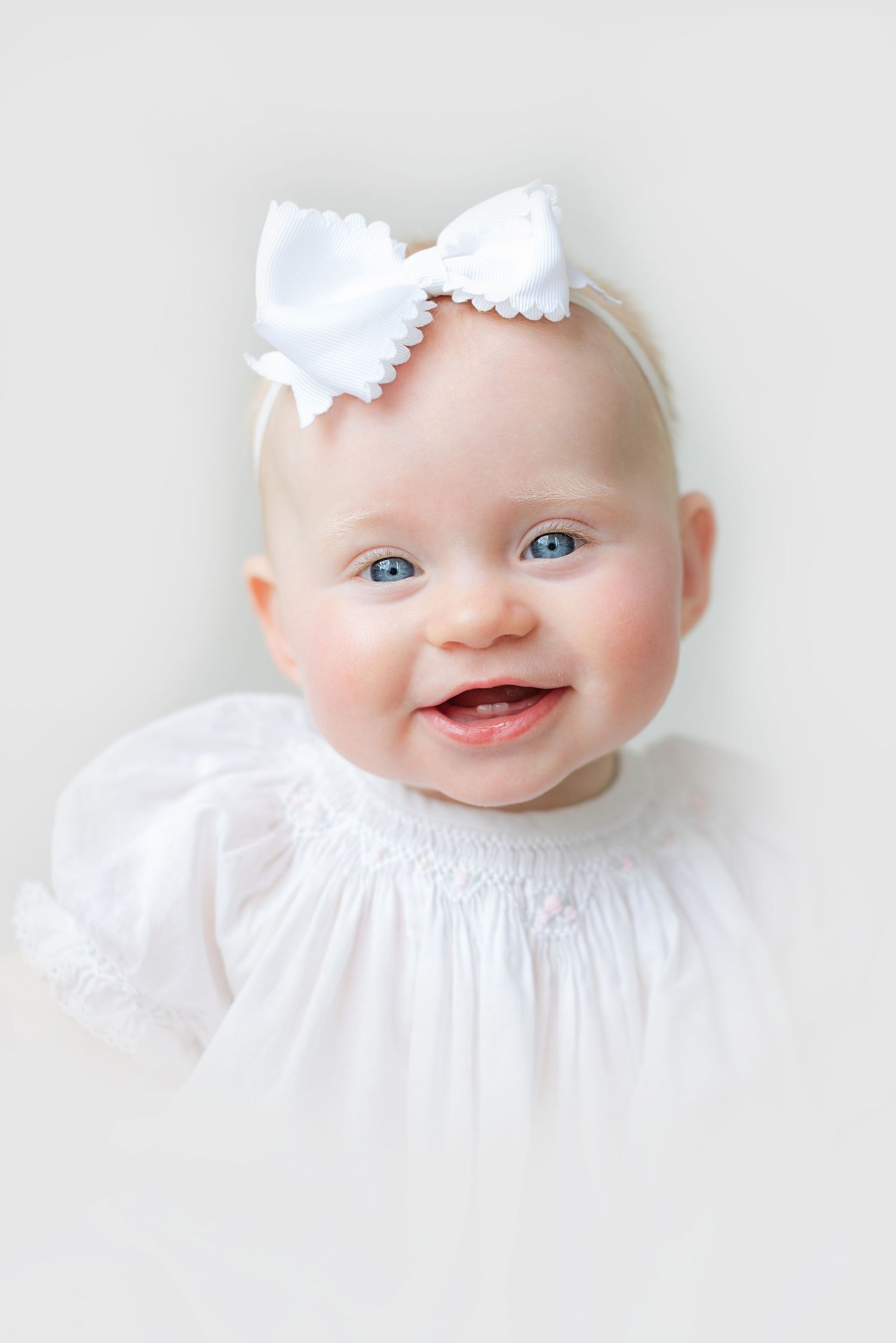 Smiling baby girl with blue eyes wearing a white smocked dress and white bow headband against a light background.
