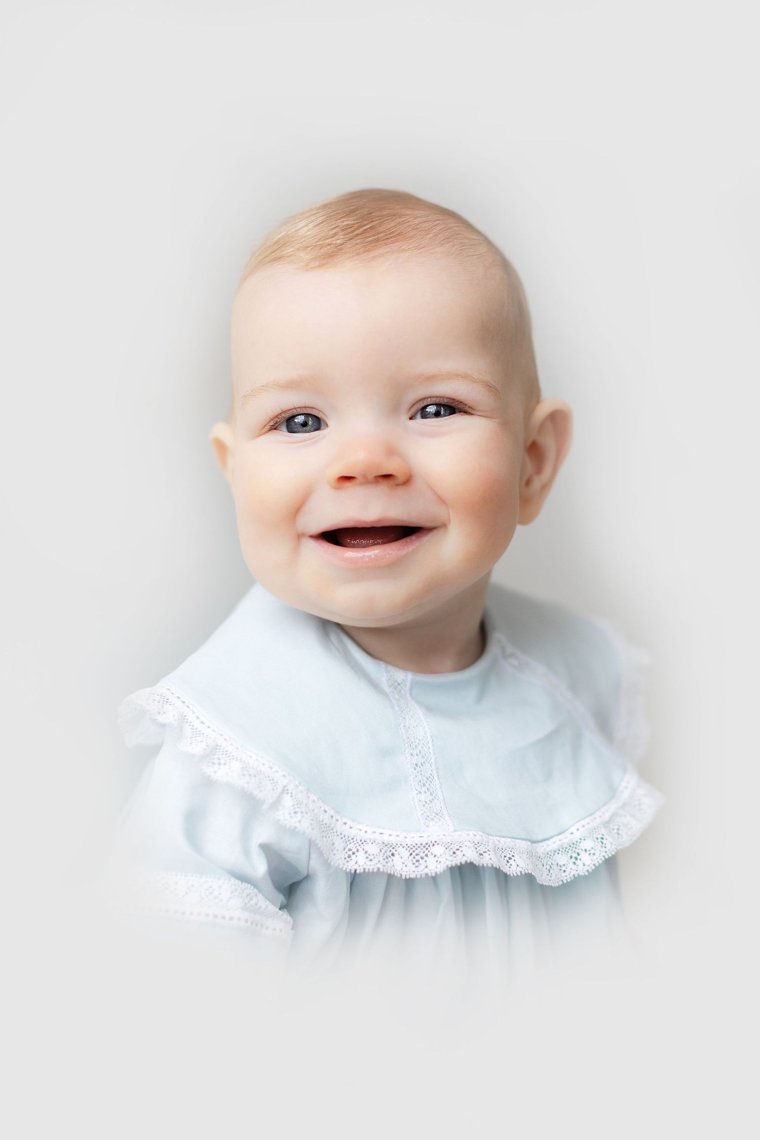 Happy smiling baby wearing a light blue lace-collar dress, looking at camera on white background.