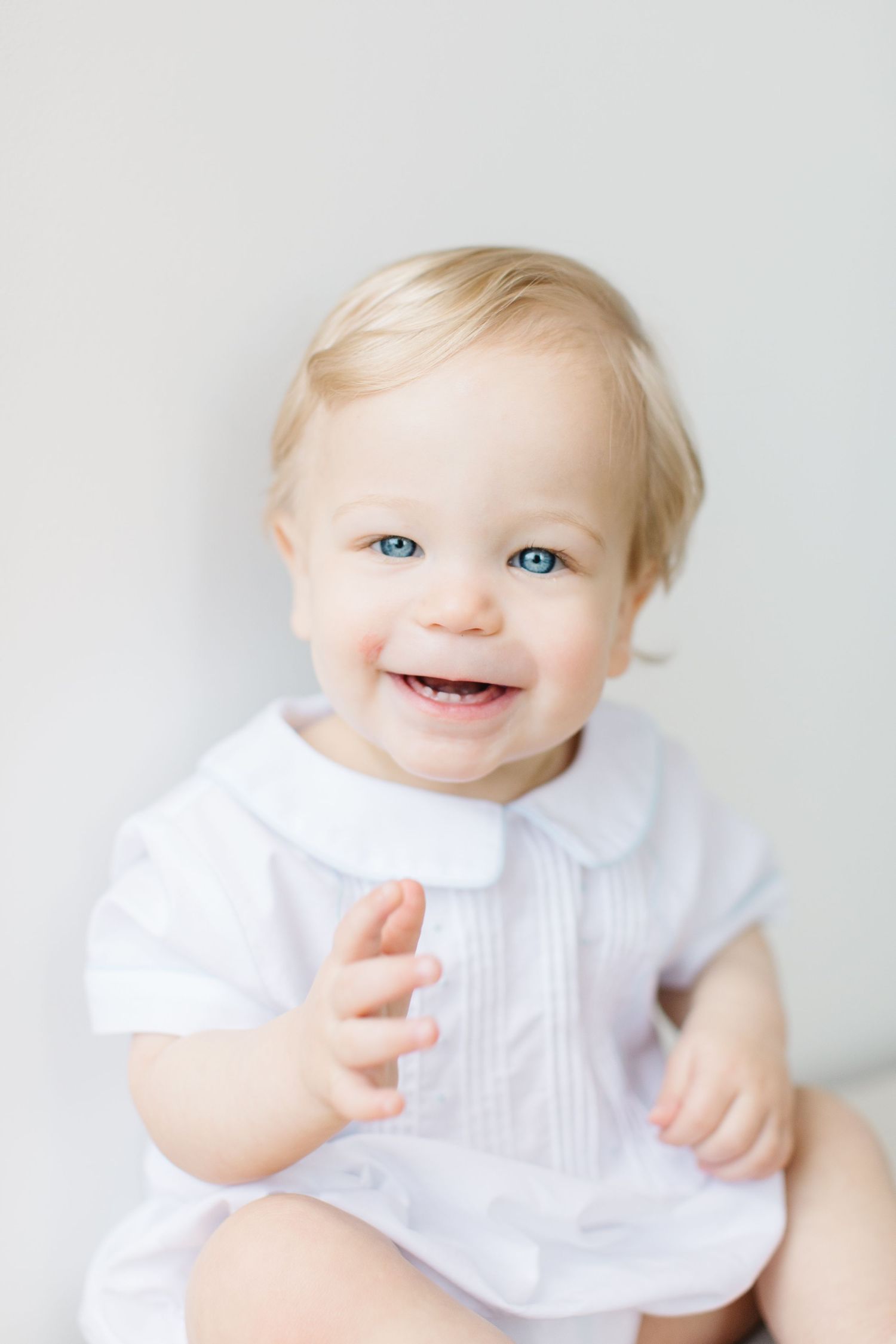 Sweet baby in white dress smiles and plays against light background in series of candid portrait photos.