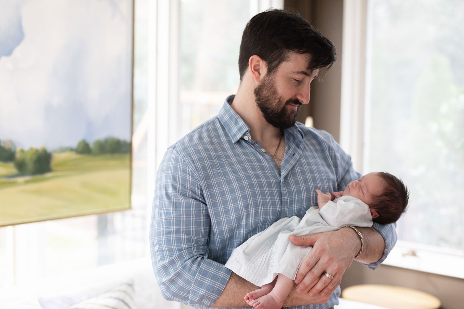 A bearded man in a checkered shirt holds a sleeping newborn baby next to a bright window.
