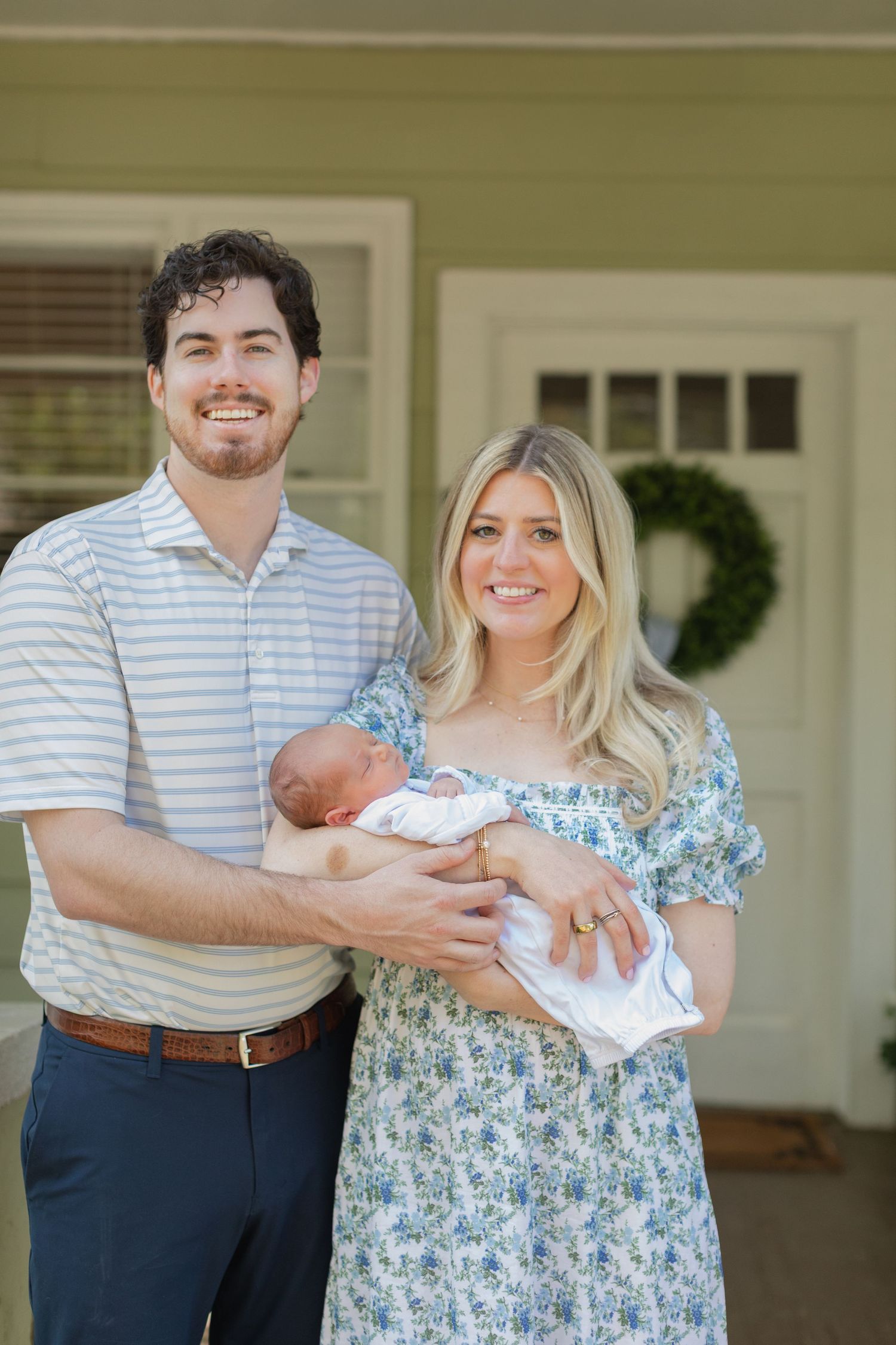 Happy couple smiling on porch holding newborn baby, man in striped shirt and woman in floral dress.
