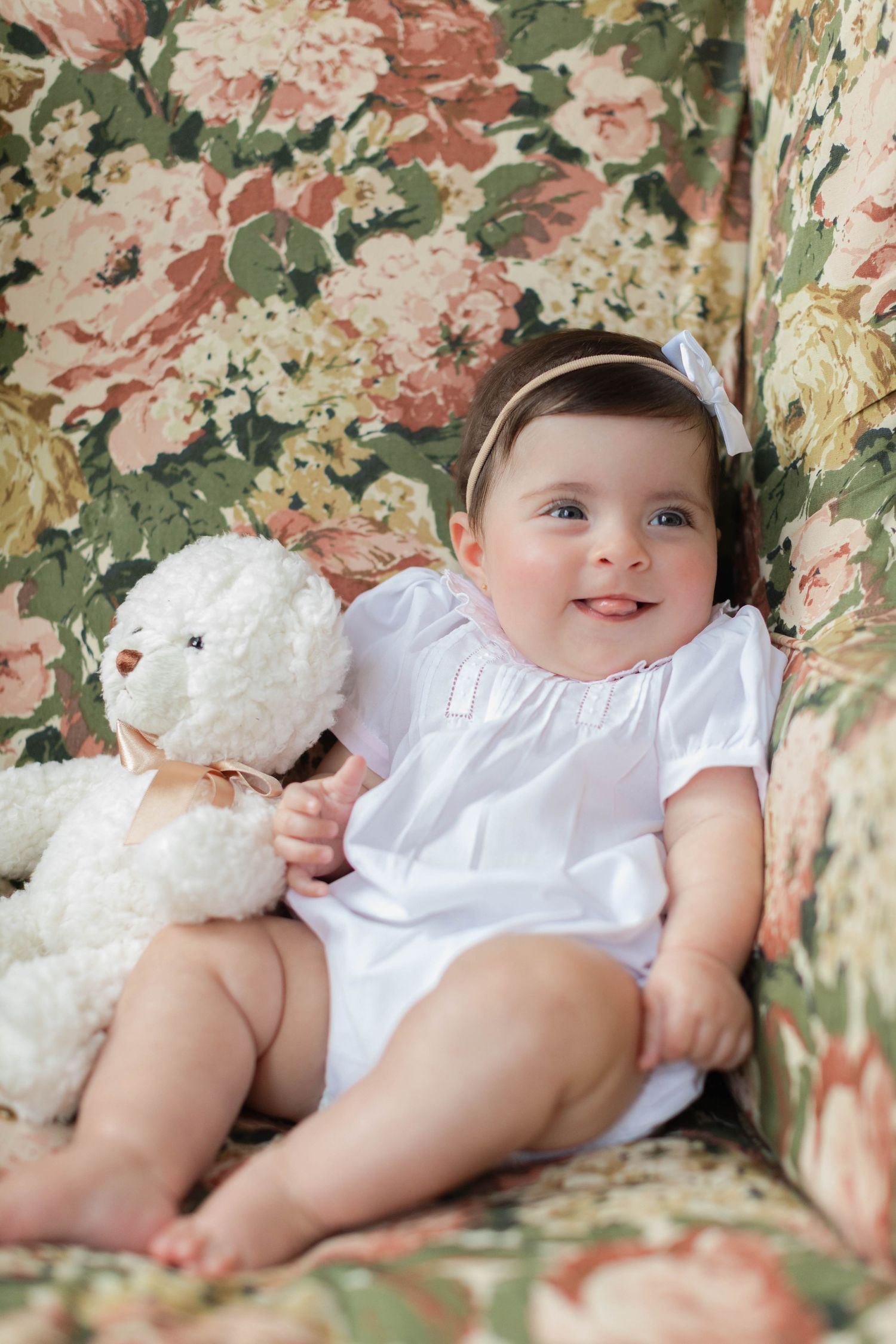 A baby in a white dress laying on floral patterned fabric with a plush teddy bear toy.