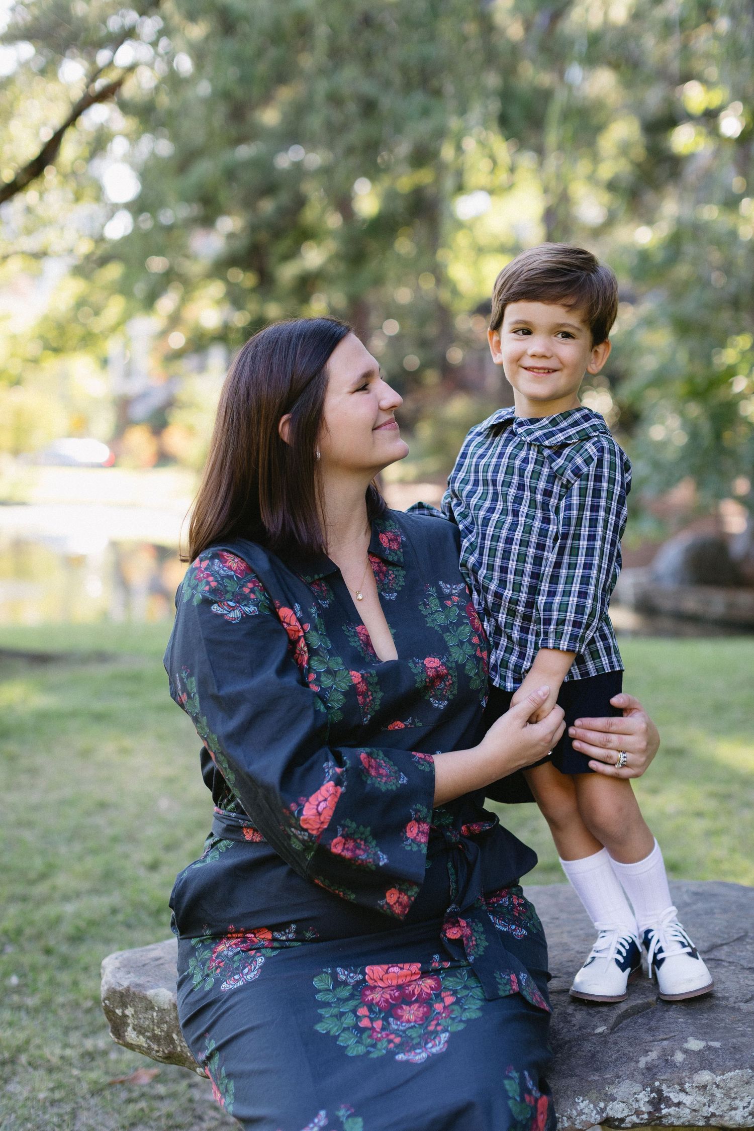 A woman in a floral dress shares a playful moment with a young child on a rock in a sunlit outdoor setting.