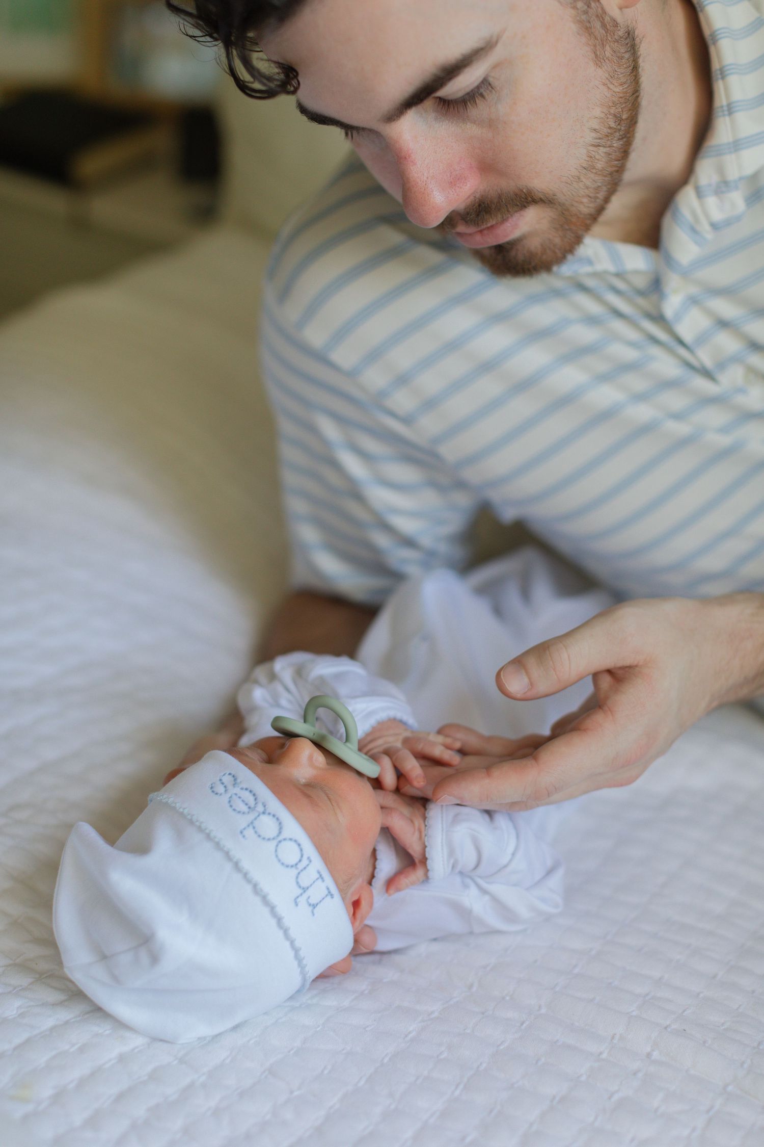 Bearded father gently holding newborn baby's hand on white quilted bed in soft natural light.