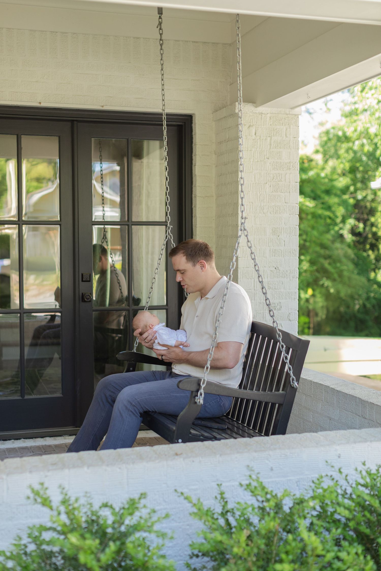 Father sitting on a porch swing holding his newborn baby outside a white brick home.