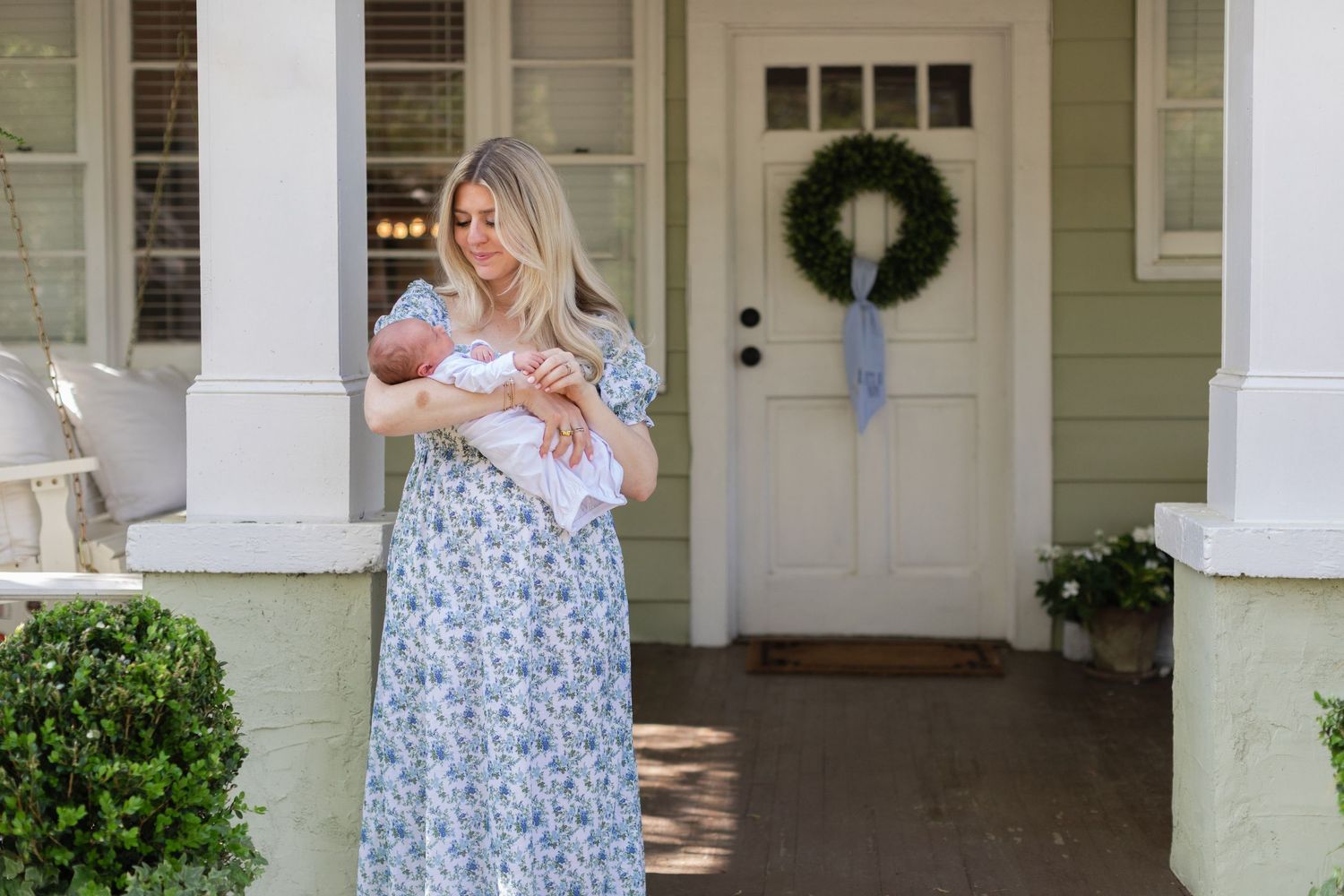 Woman in floral maxi dress holding newborn baby on front porch with wreath-decorated door.