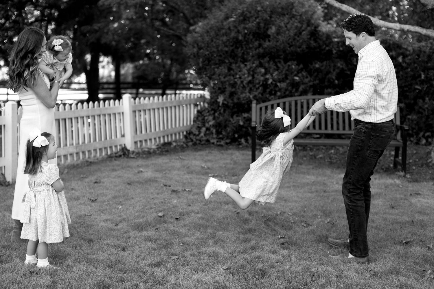 Black and white photo of playful outdoor scene with someone being swung around by an adult in a backyard with picket fence.