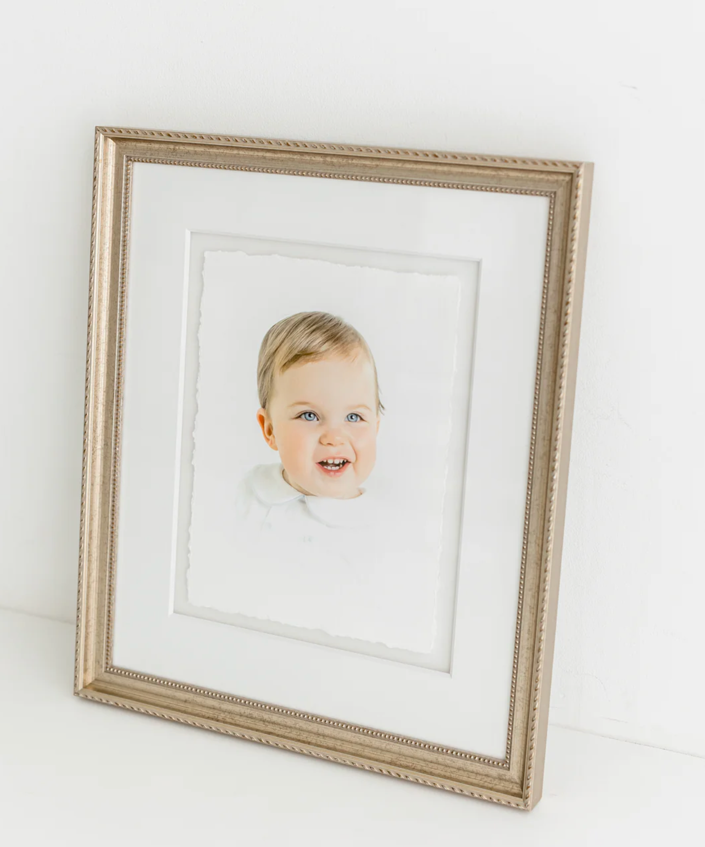 A framed portrait of a smiling toddler with blue eyes displayed in an elegant silver frame with white matting.