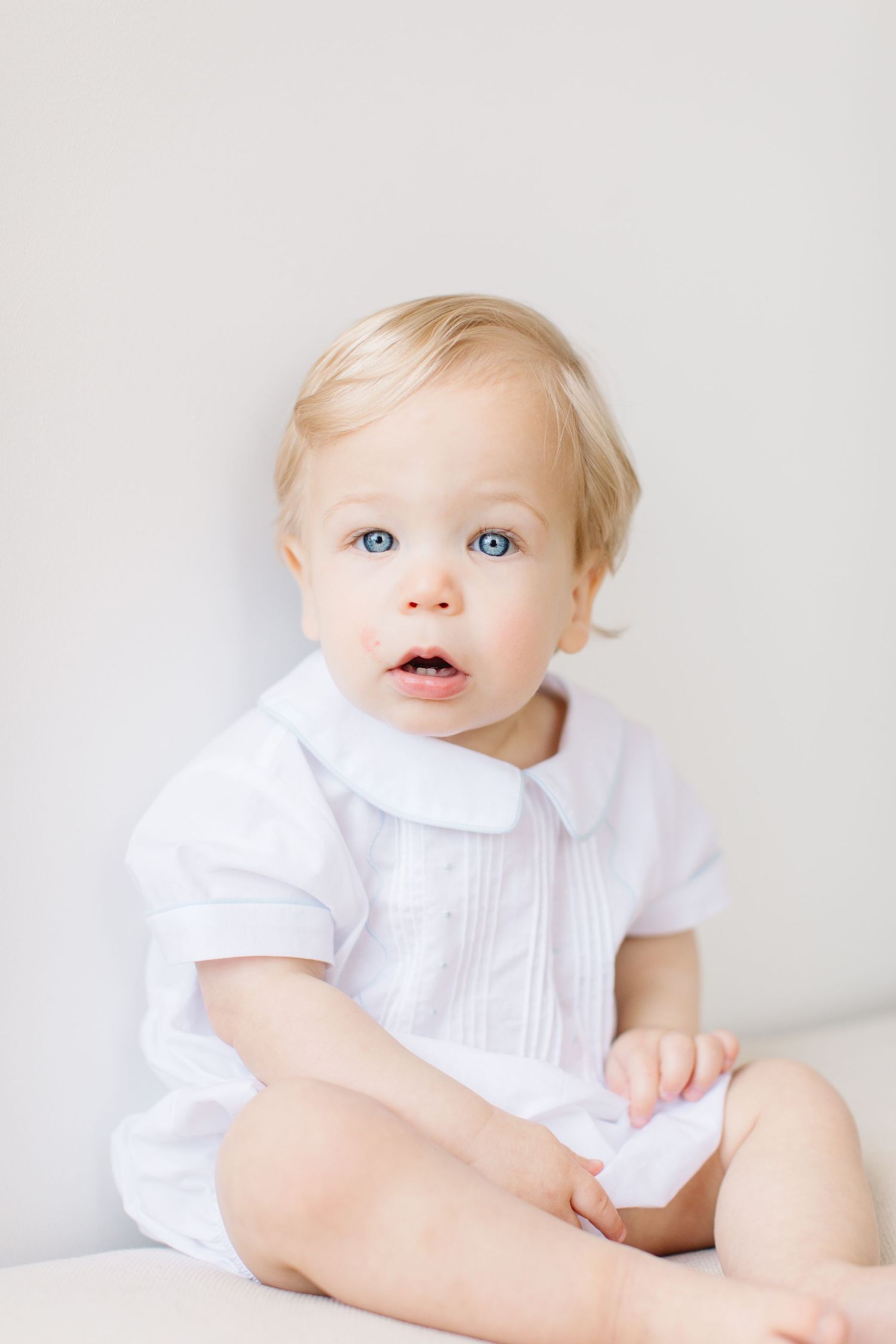 Baby in white dress sitting and looking up with bright blue eyes against neutral background.
