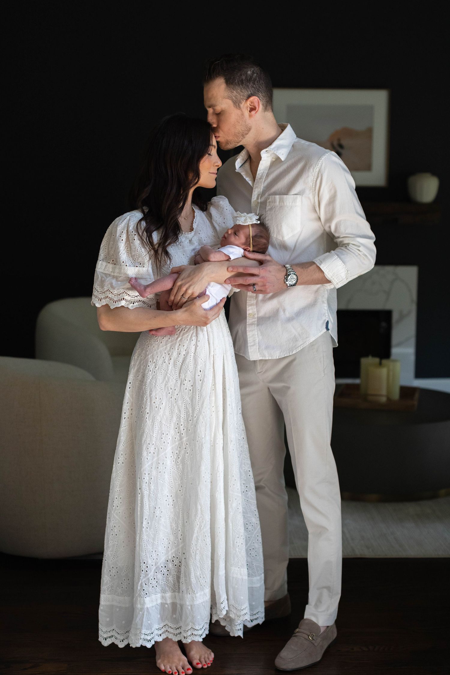 Couple in white attire shares intimate moment while holding newborn baby in dark room.