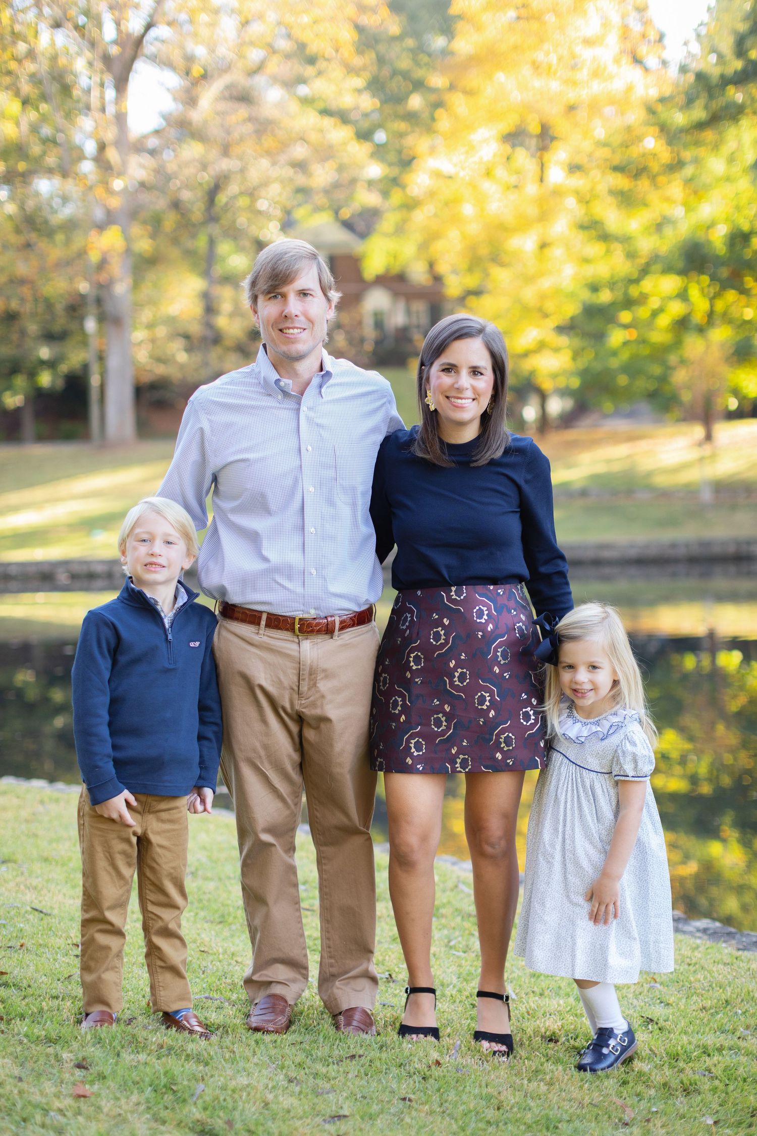 A family of four poses together on a grassy hill during autumn with golden foliage in the background.