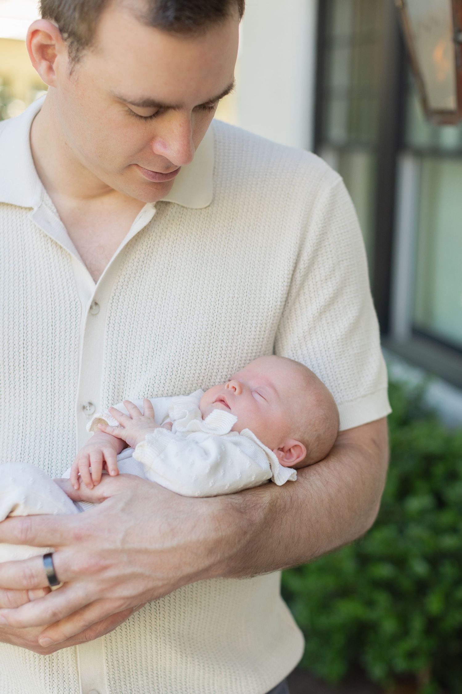 Father in cream shirt tenderly cradling sleeping newborn baby outdoors near home entrance.