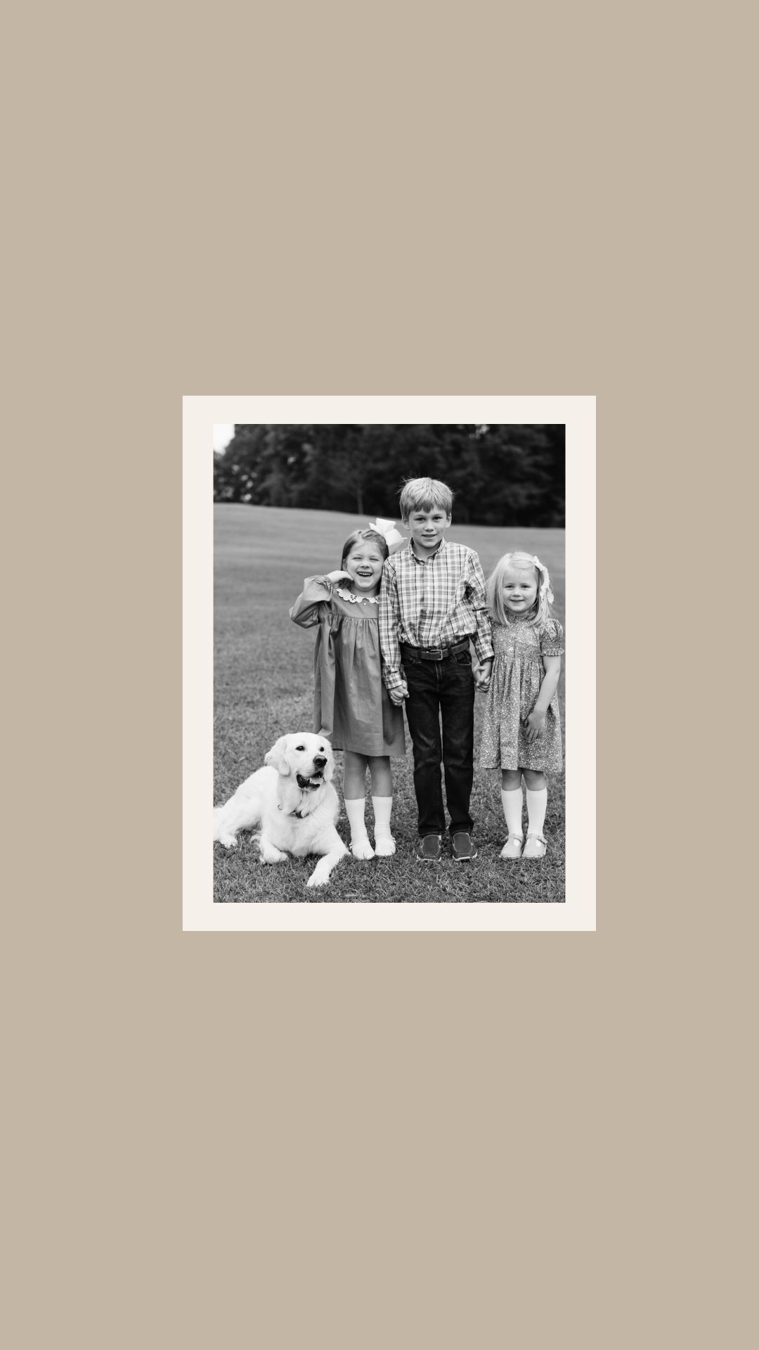 A black and white portrait of three young children and a white dog posed together outdoors.