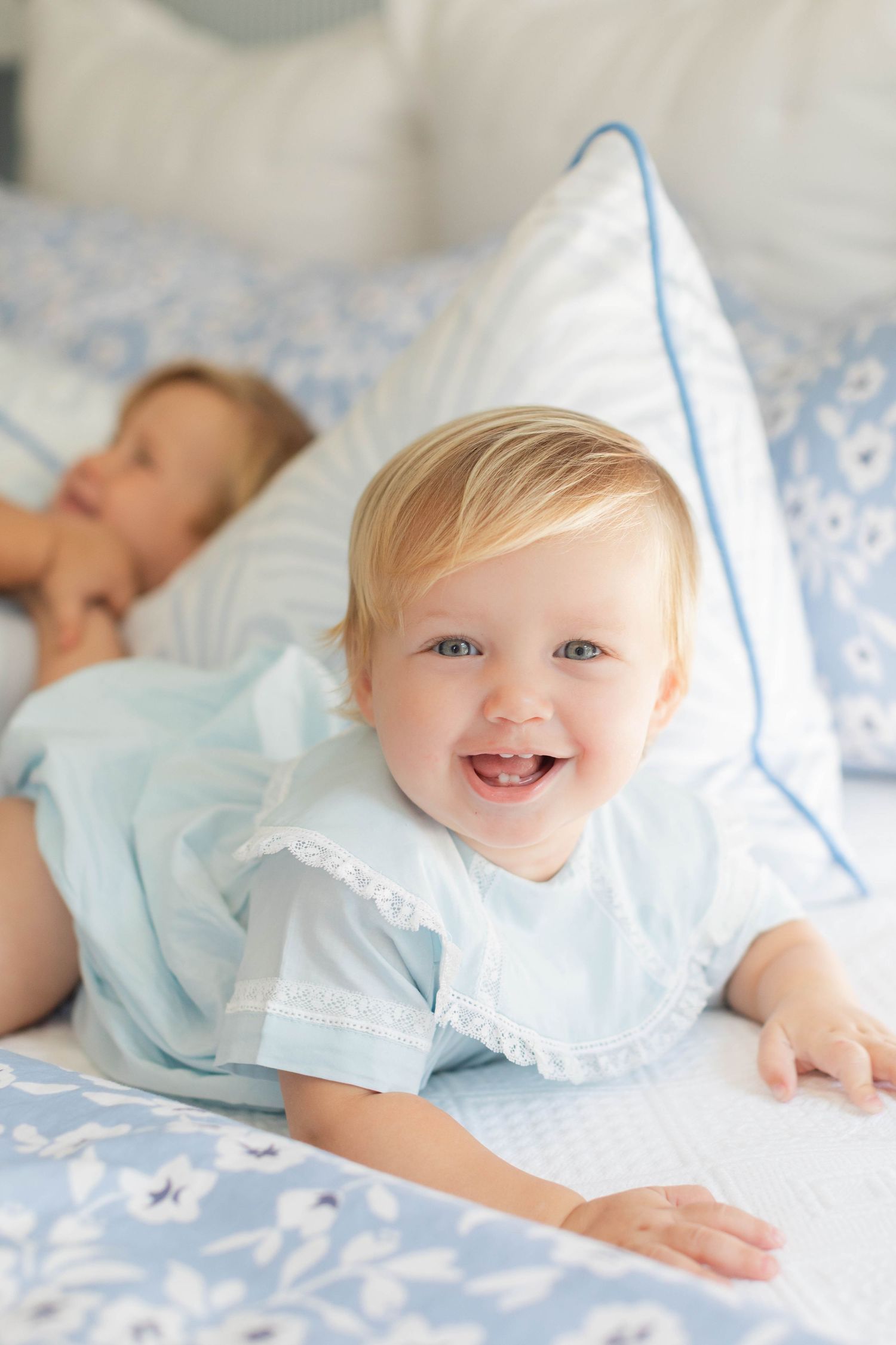 Smiling baby wearing light blue dress lying on blue and white patterned bedding next to sleeping figure.