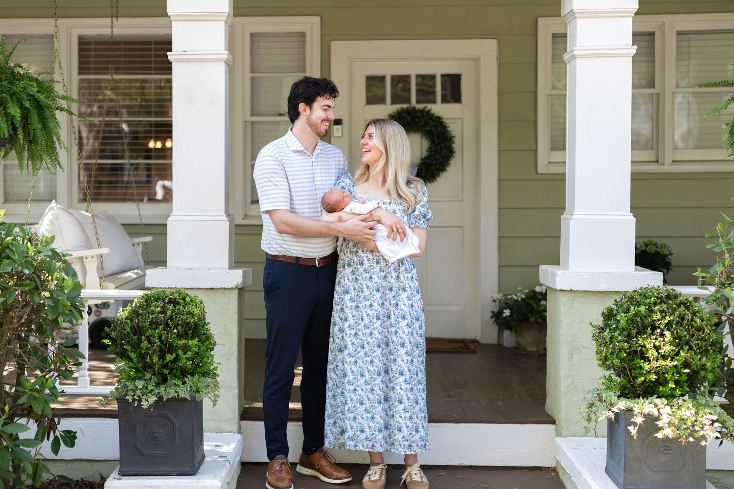 A couple stands on a charming green home's porch, smiling while holding their newborn baby in a floral dress.