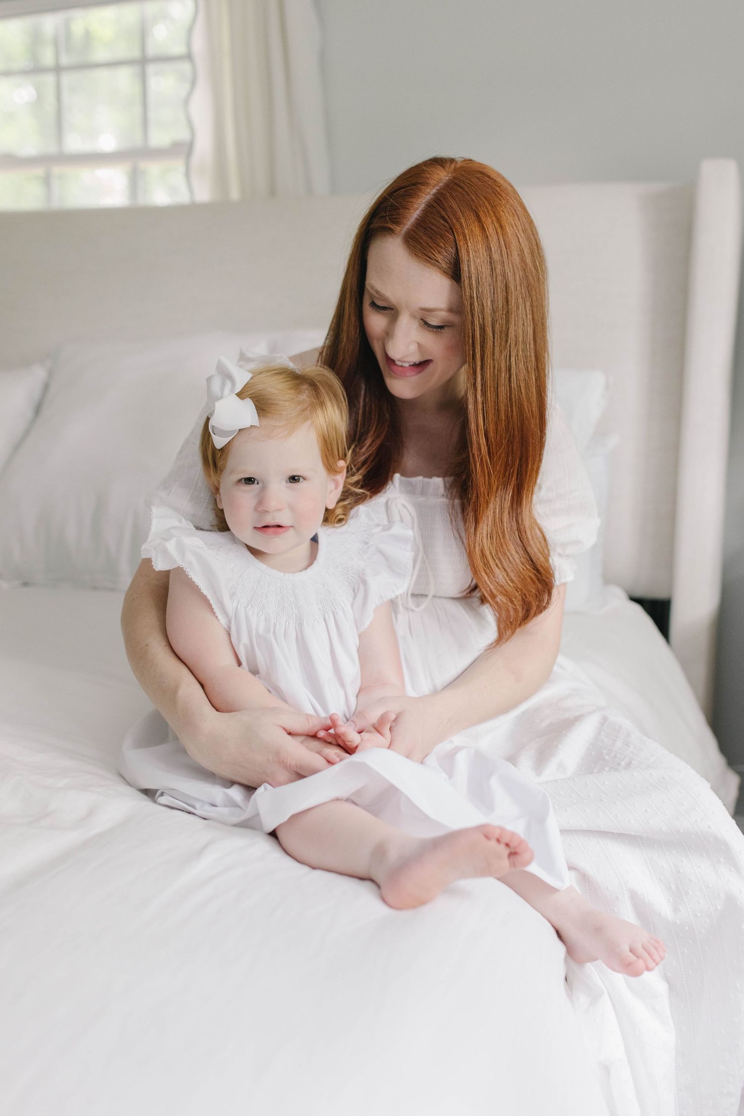Mother and child wearing white dresses sit together on a bed with crisp white linens.