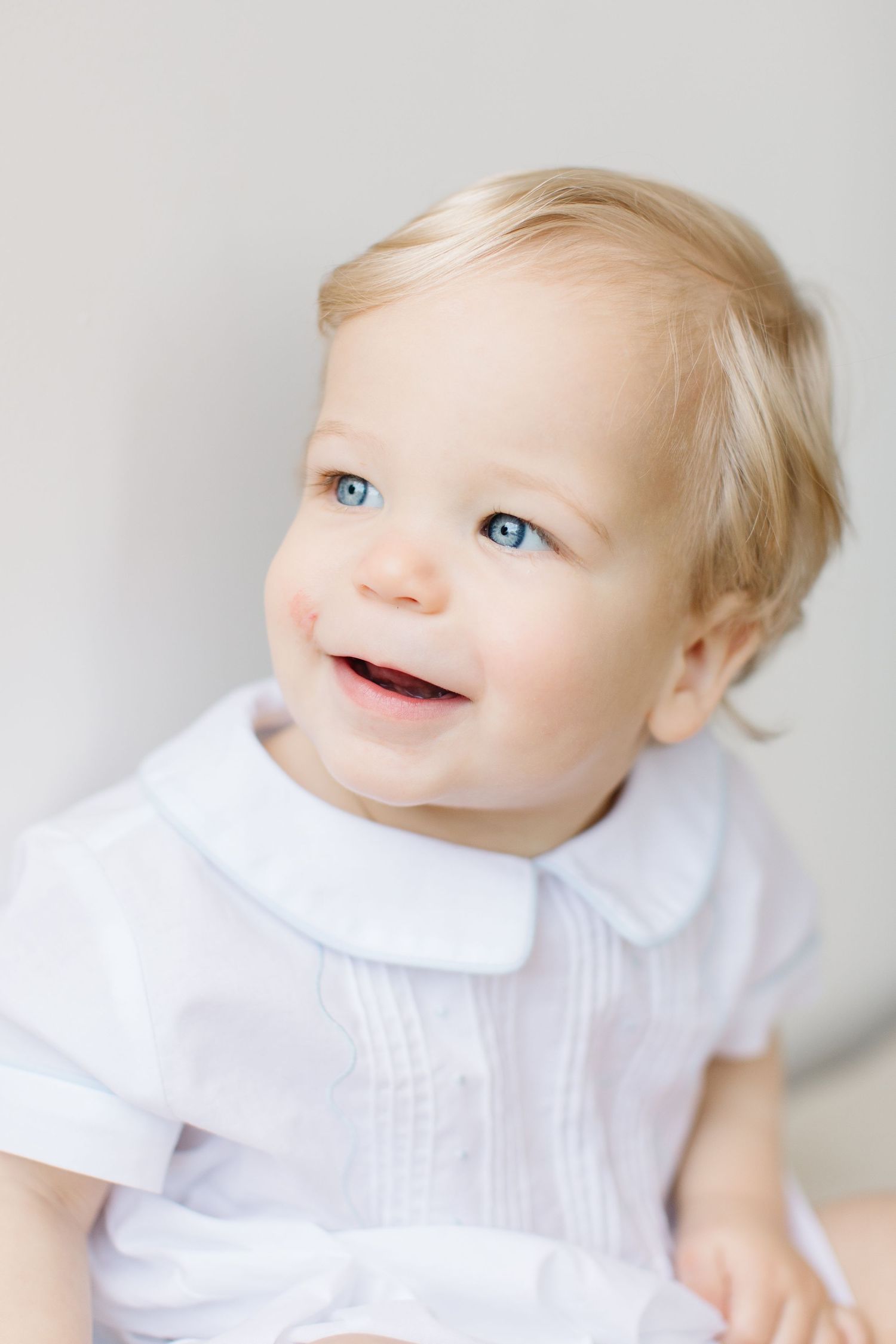 Bright-eyed baby in white outfit smiles and looks up against light background in portrait series.