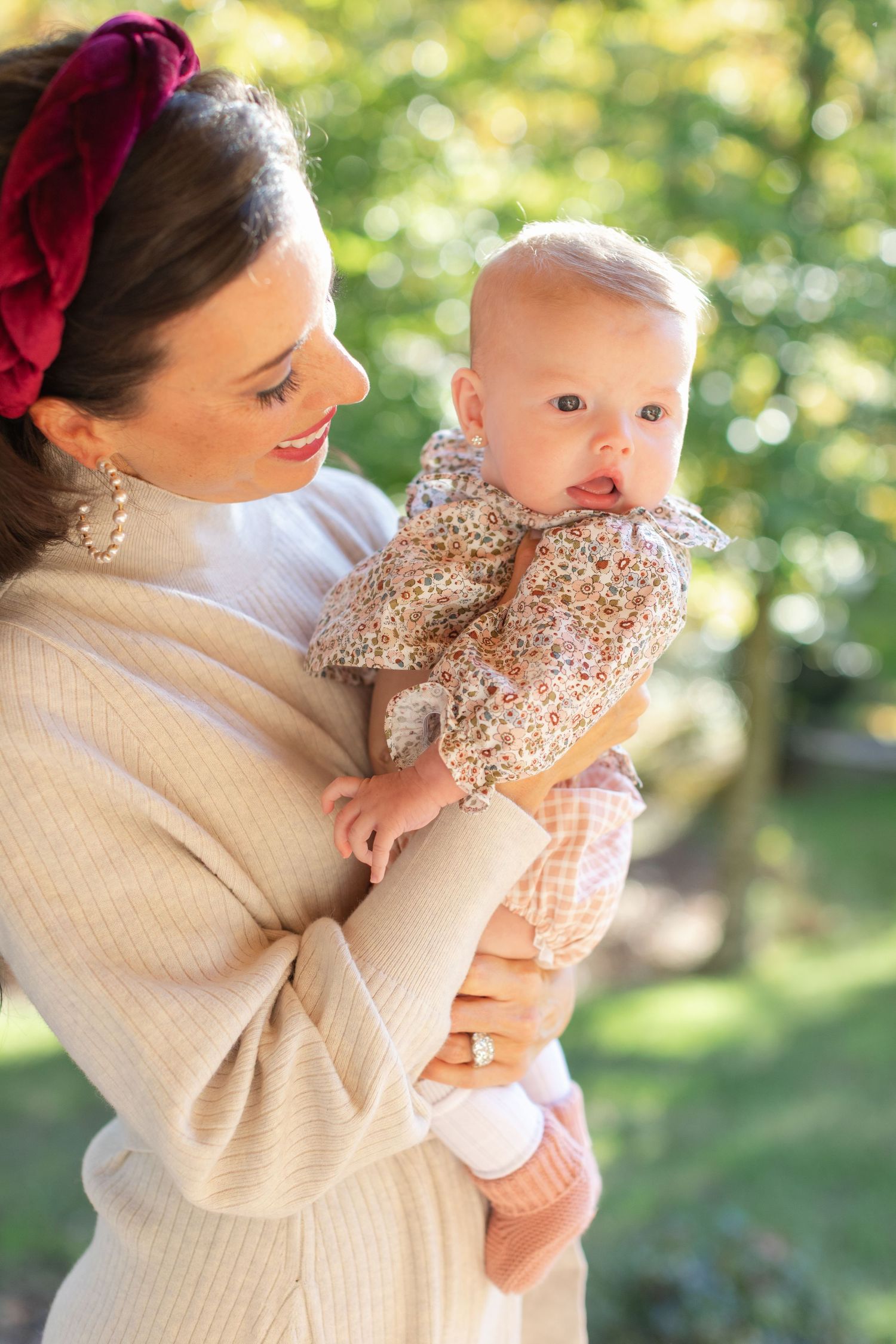 Parent in cream sweater holds smiling baby wearing floral outfit against blurred outdoor background.