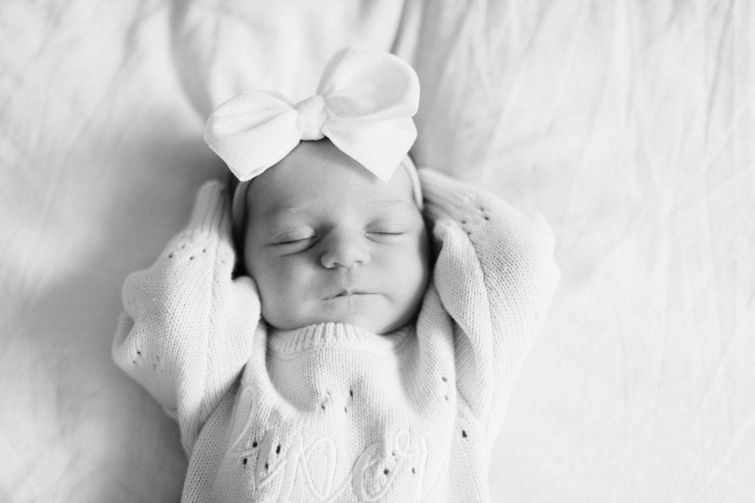 Black and white photo of a sleeping newborn baby wearing a white knit hat and swaddled in soft fabric.
