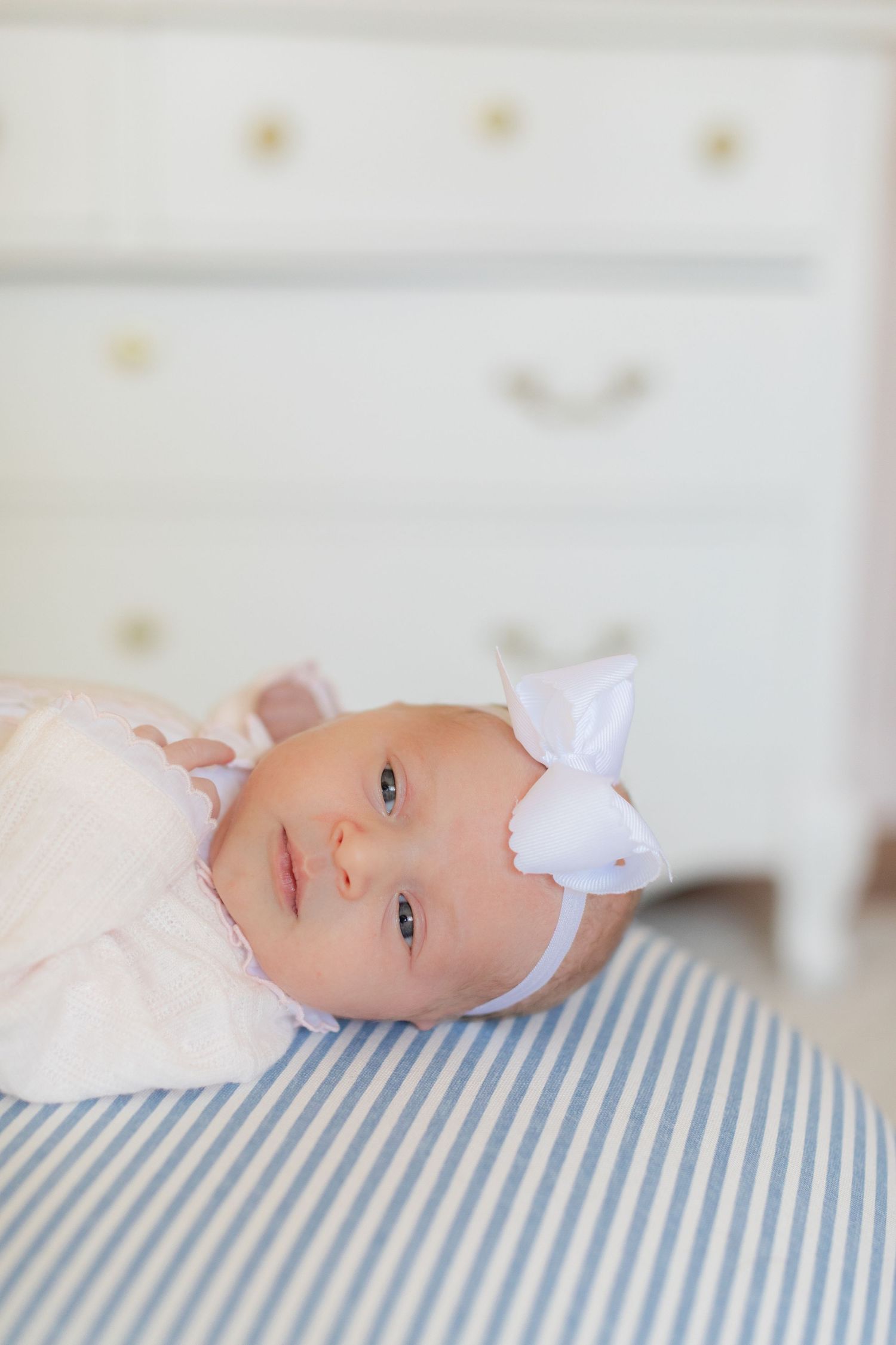 Infant with white bow headband lying on blue striped surface near white furniture.