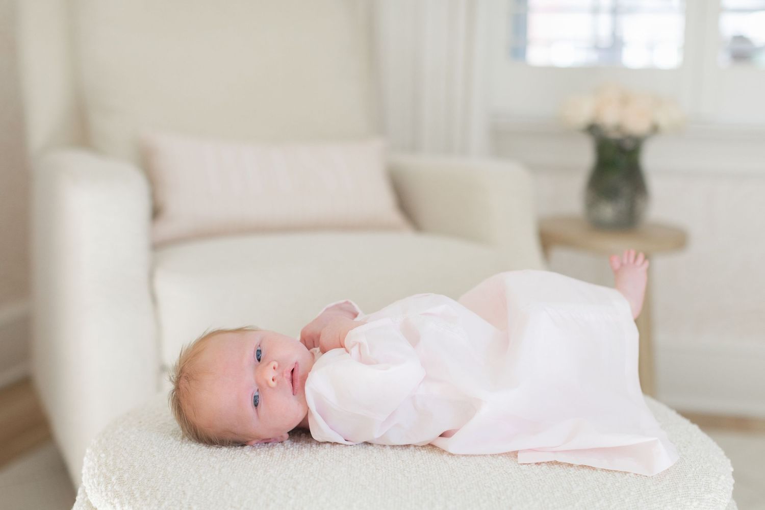A baby wearing white sleepwear lies on a cream-colored couch in a bright, airy room with soft decor.