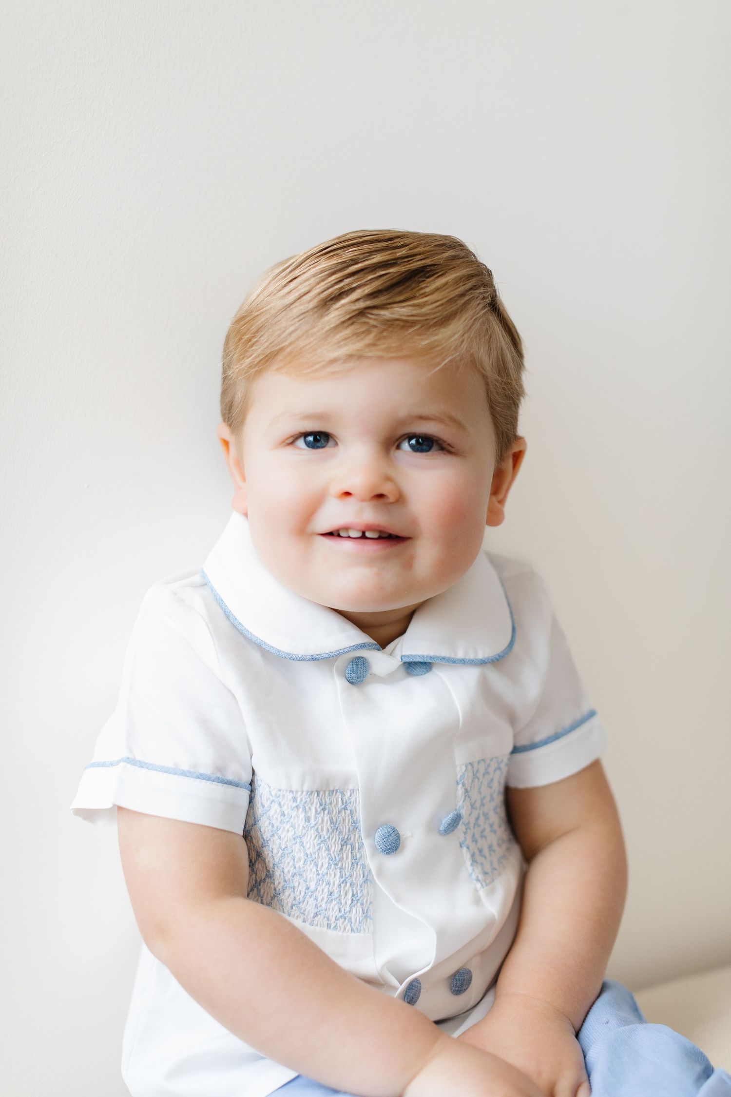 A young child in a white and blue smocked outfit smiles while sitting against a light background.