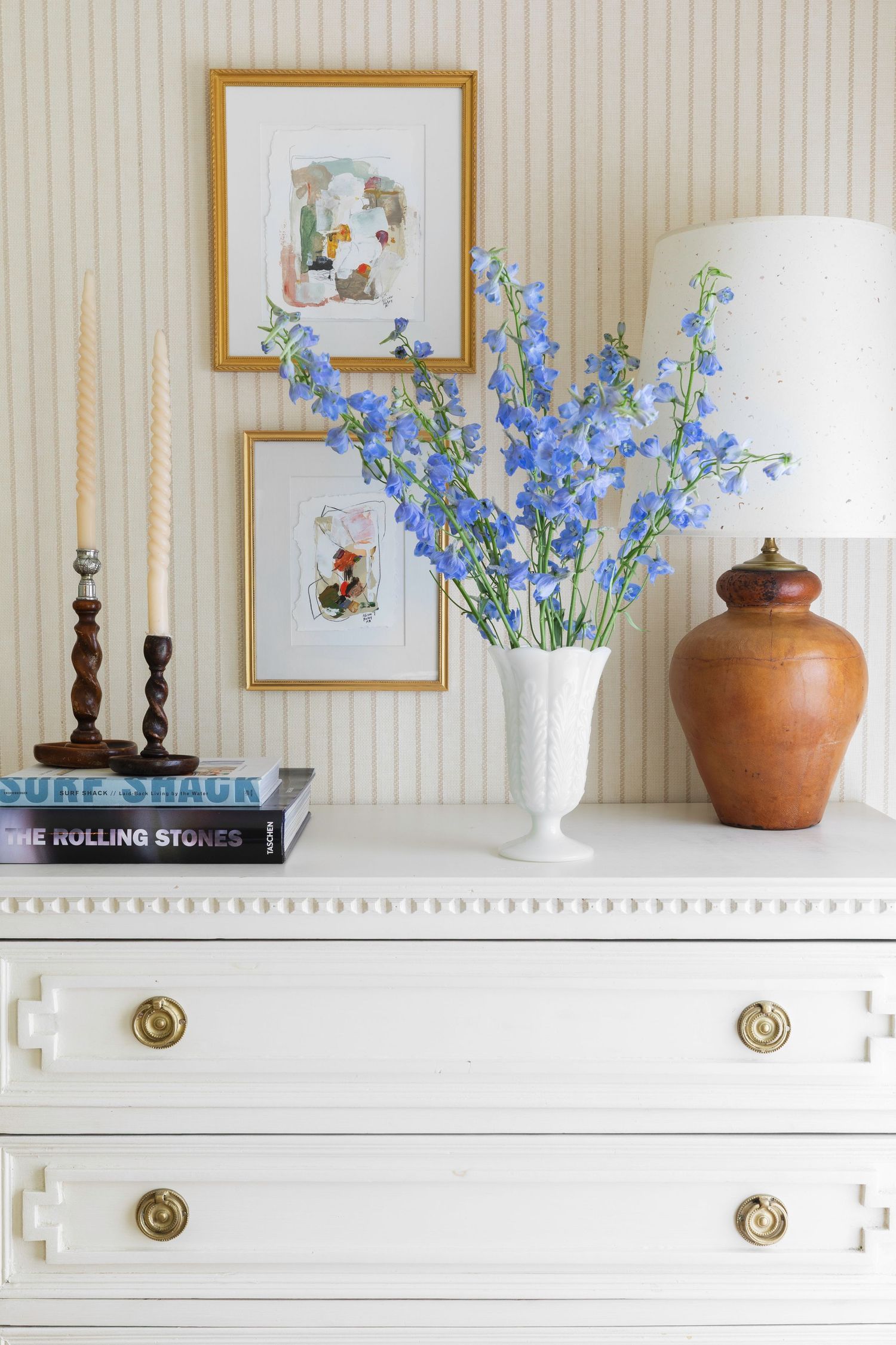 White dresser with brass hardware topped with blue delphinium flowers, framed artwork, candles, books, and a wooden lamp.