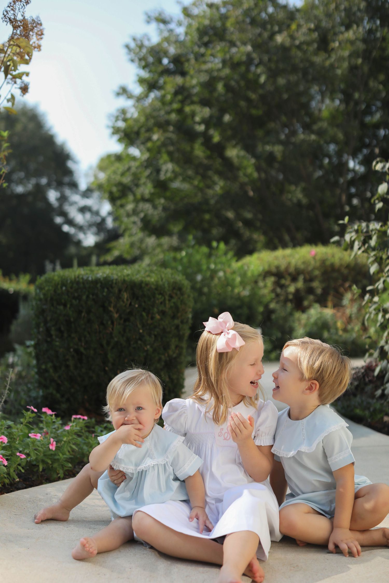 Children lay down in a row on a garden path, laughing and playing together in an outdoor setting.