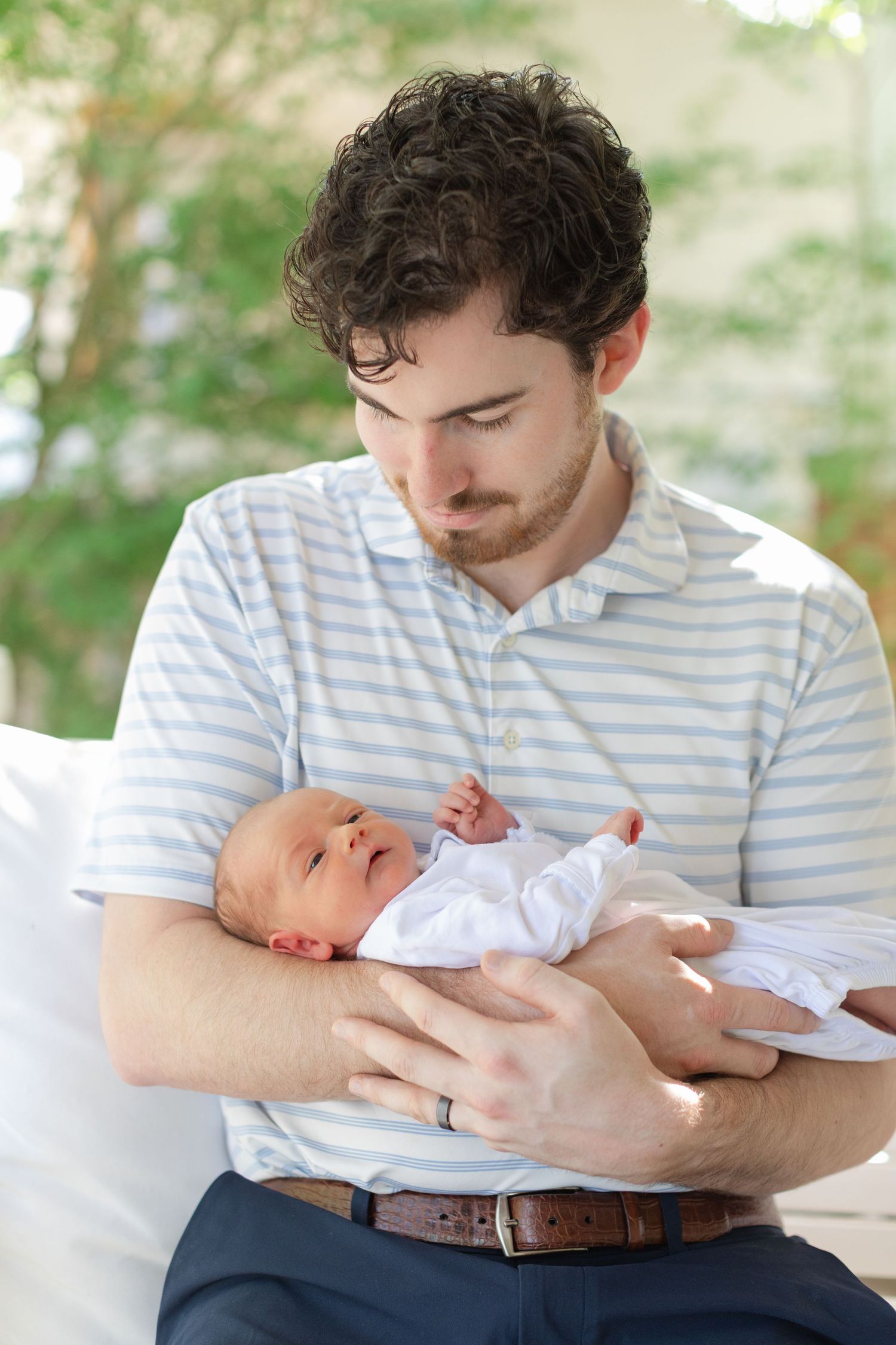 Young father in striped polo shirt tenderly cradling newborn baby outdoors on white cushioned seat.
