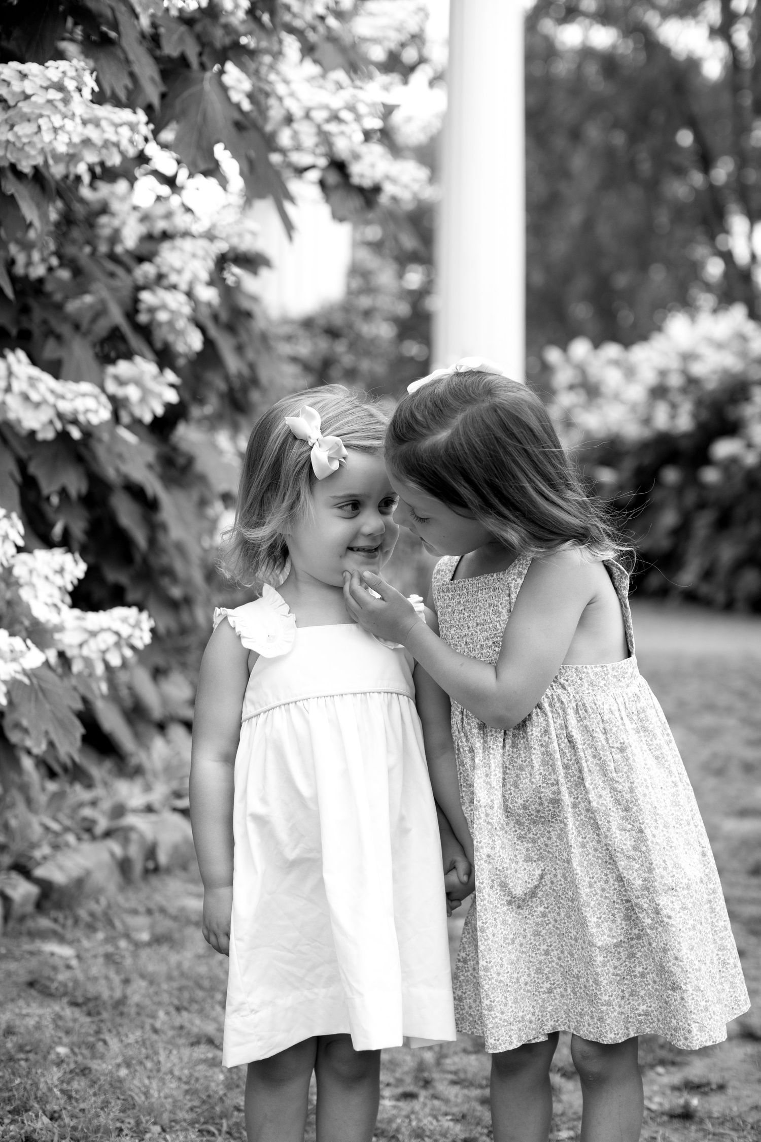 Two young girls in dresses share a sweet whispered secret outdoors near blooming flowers in black and white photo.